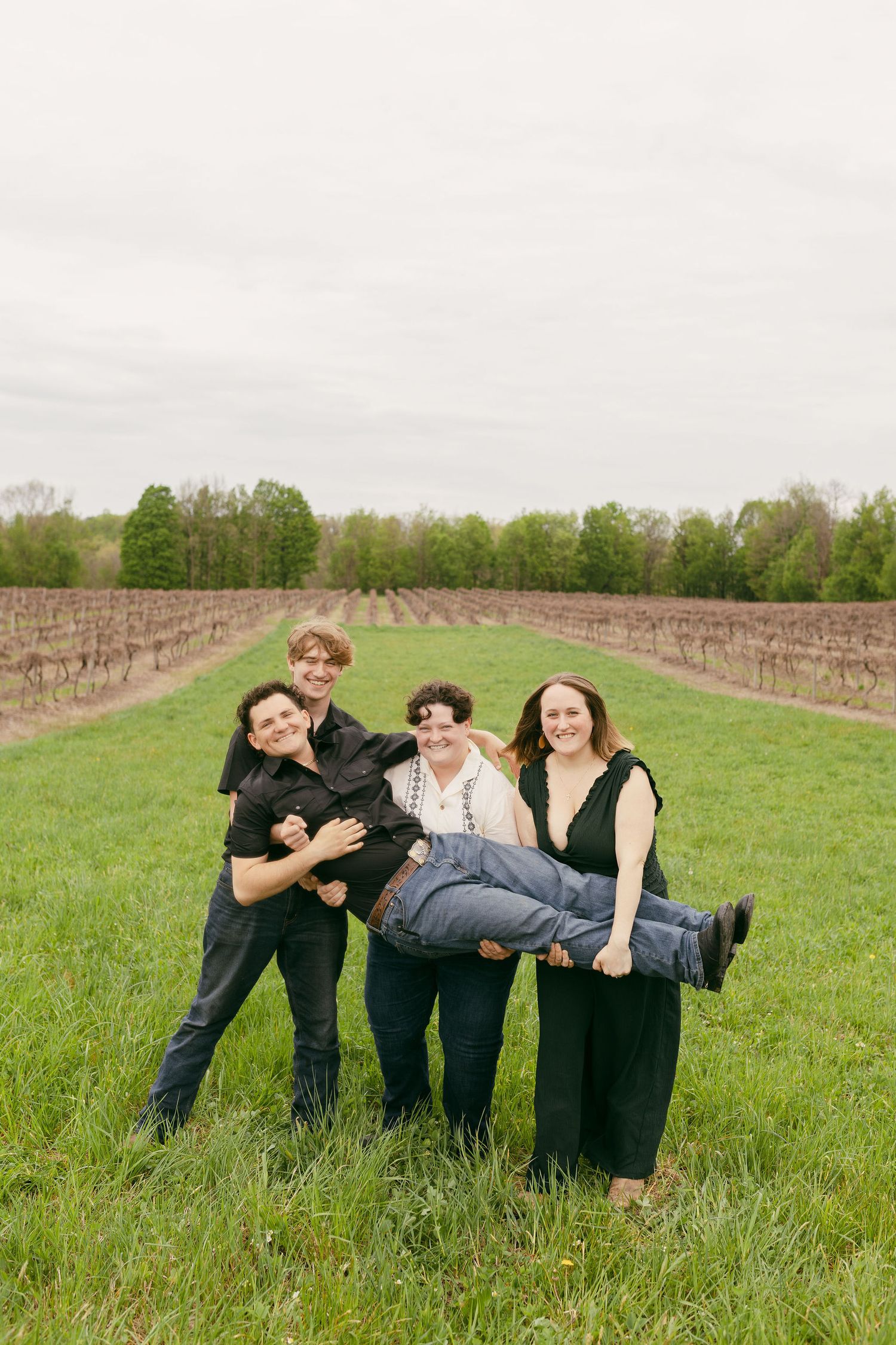 People in dark clothing playfully carry someone across a green spring field.