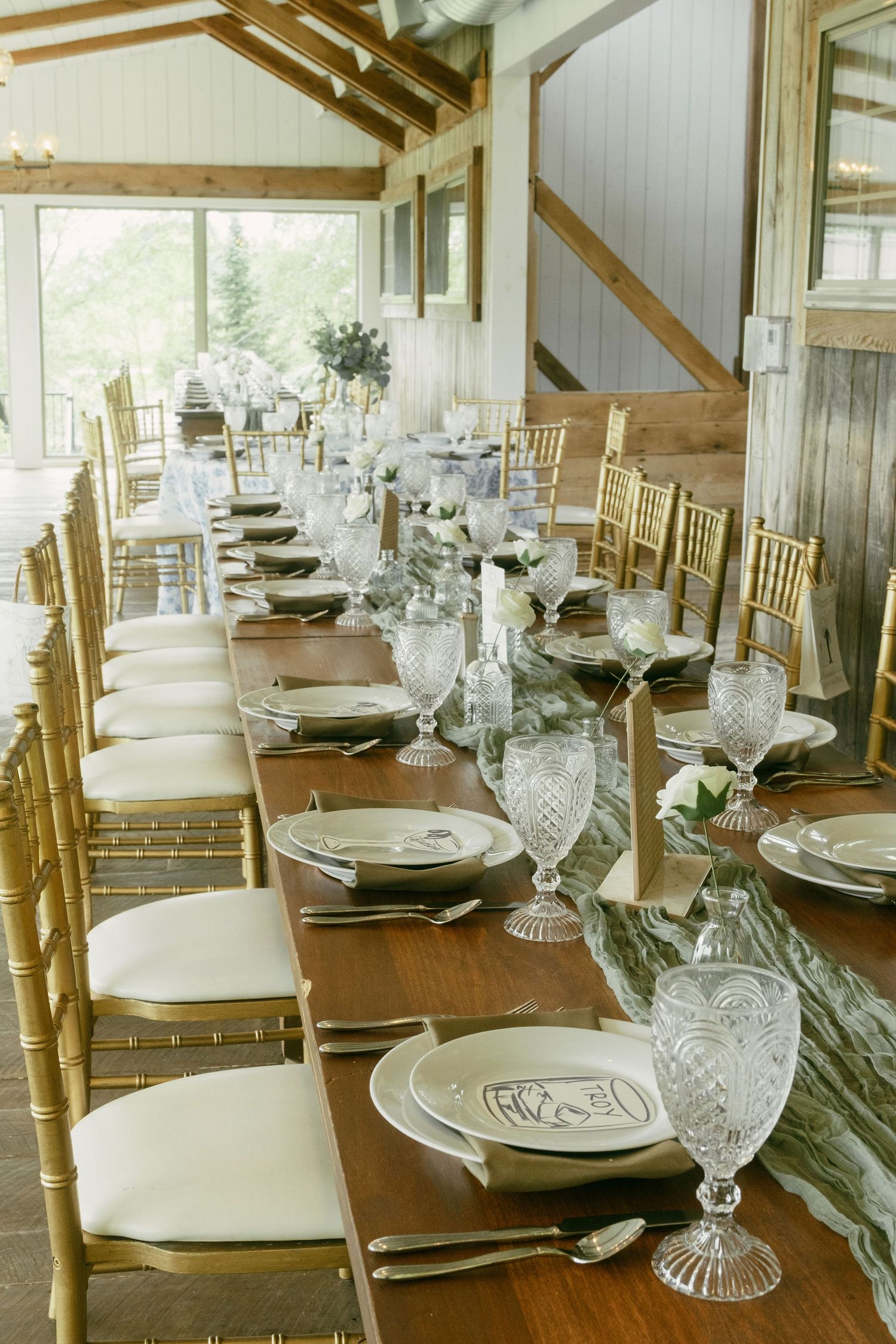 Elegant rustic wedding reception table with gold chairs and crystal glassware in a barn setting.