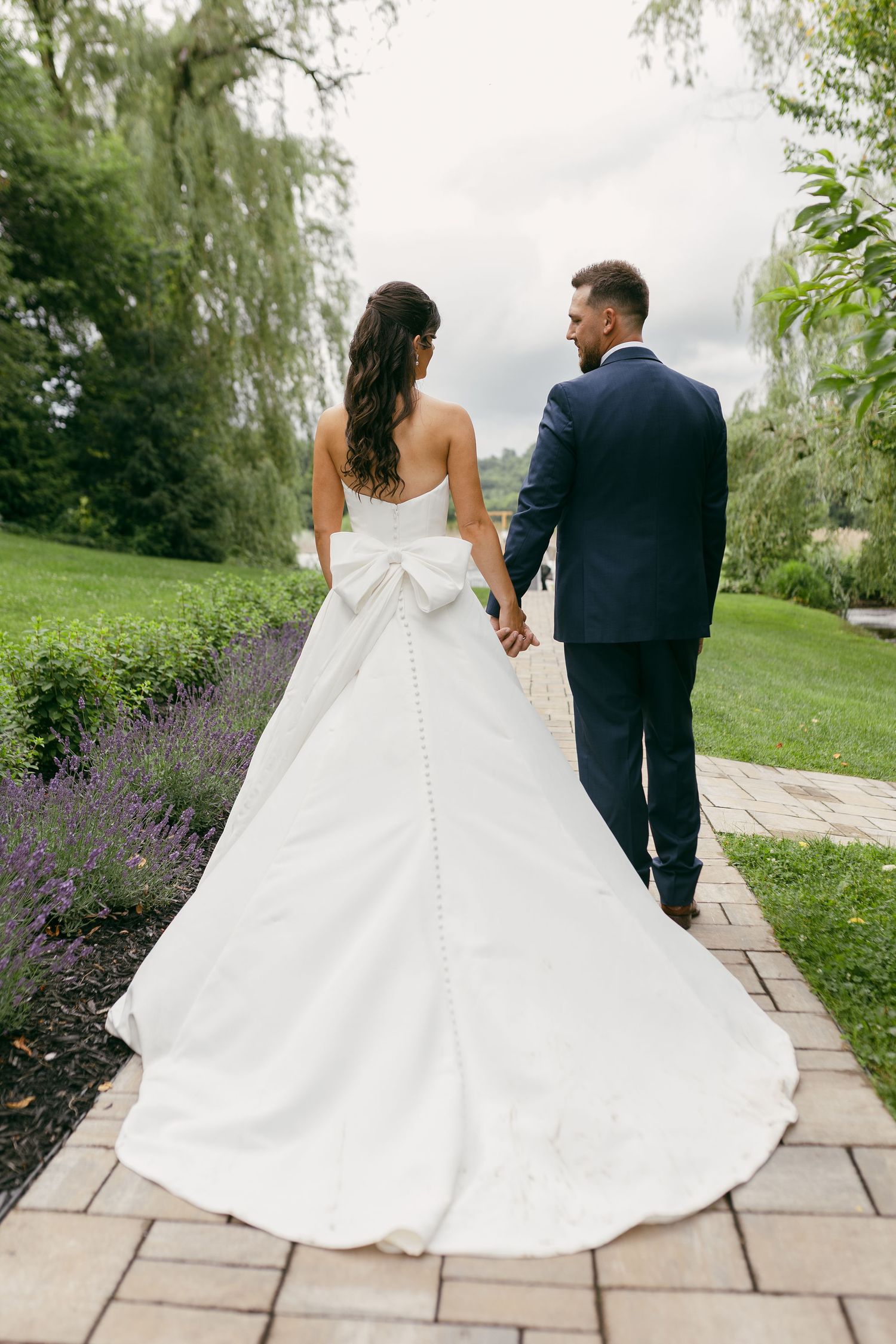 Couple walks hand in hand along stone garden path with flowing white dress.
