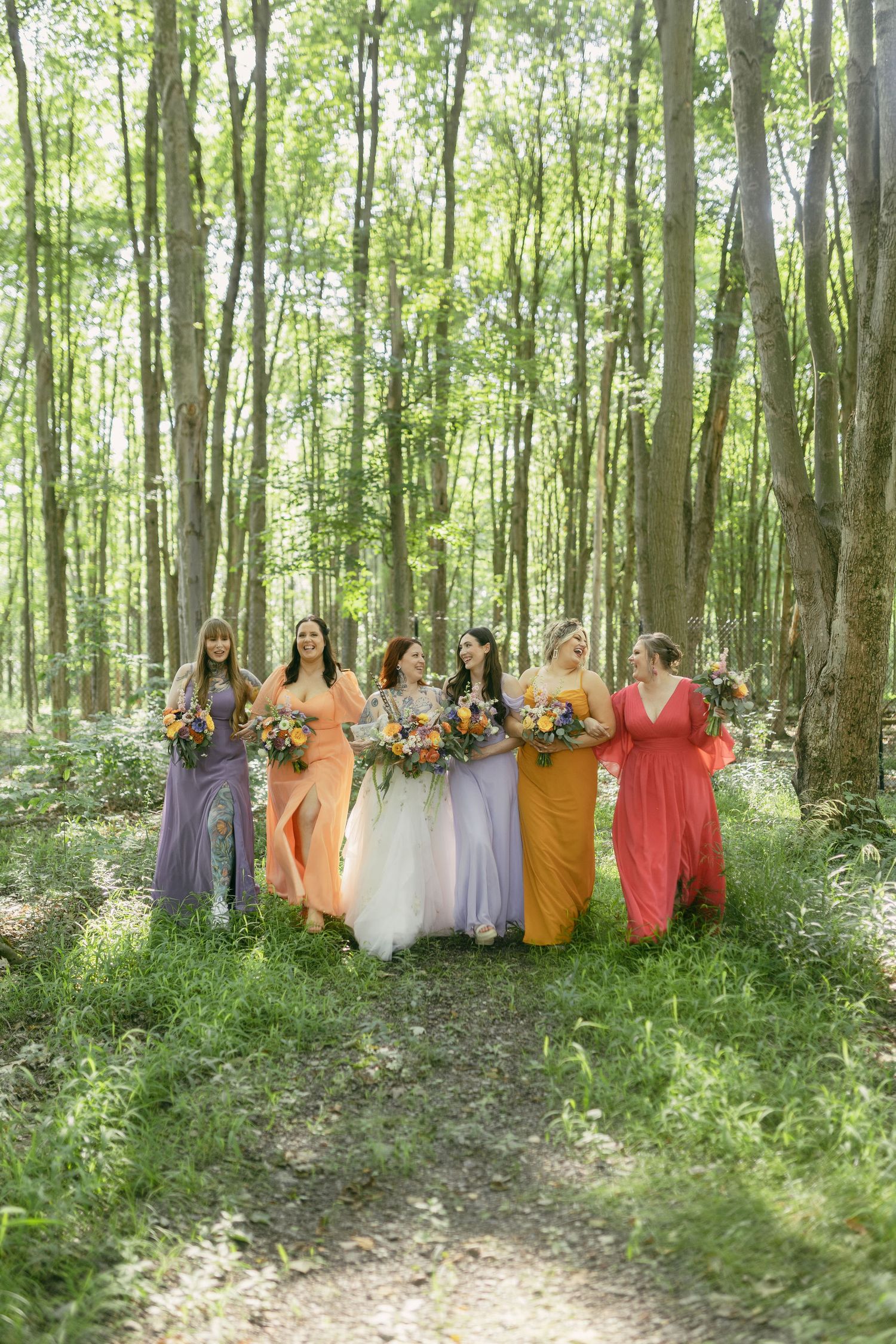 Wedding party wearing vibrant dresses stands together in a wooded area holding floral bouquets.