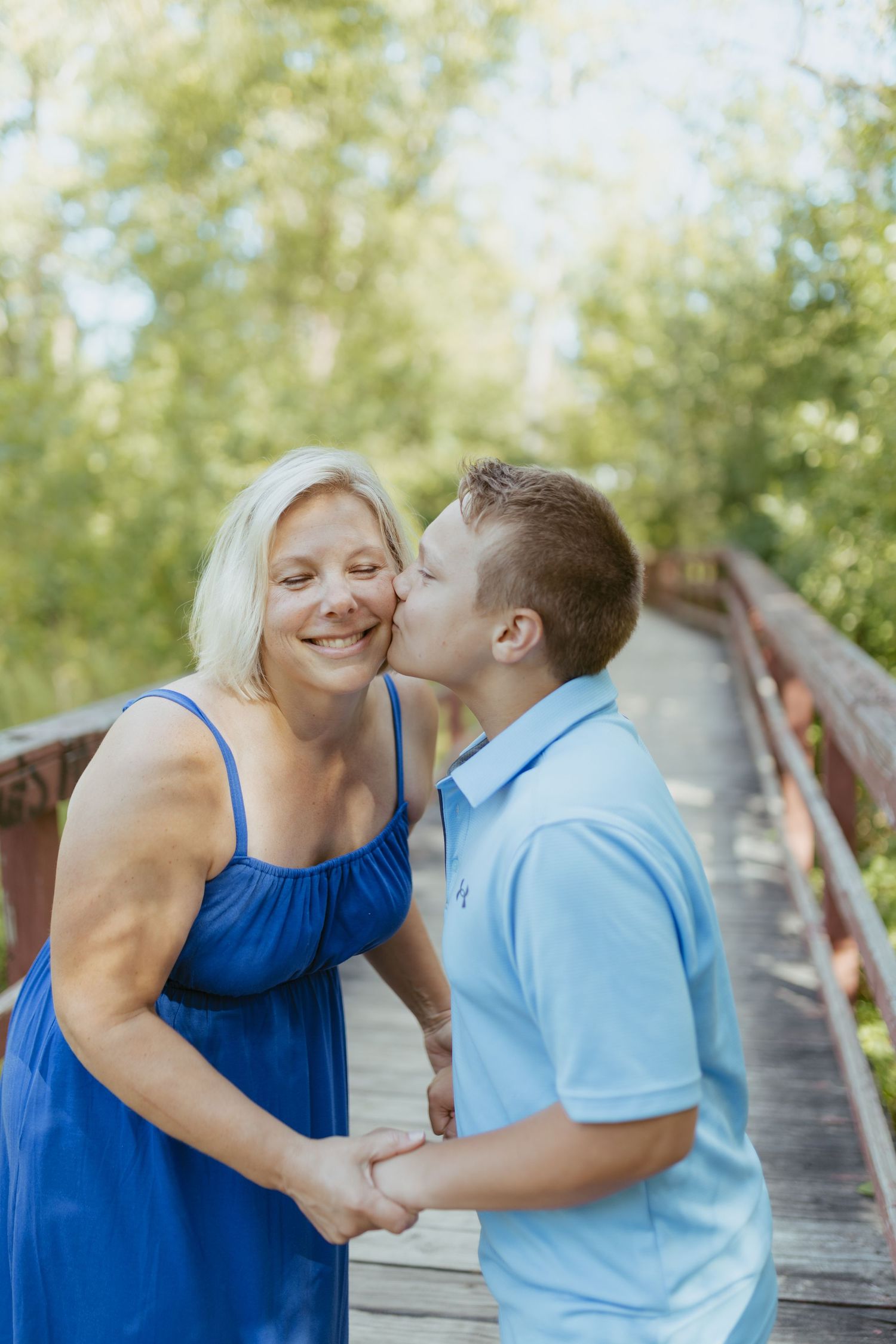 A teenage boy in a light blue polo shirt kisses his smiling blonde mother in a blue dress on a wooden bridge.
