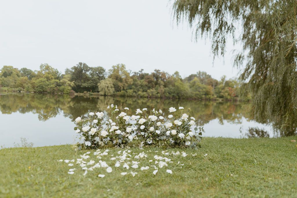 White floral arrangement on green grass beside a serene lake with weeping willow and autumn trees backdrop.