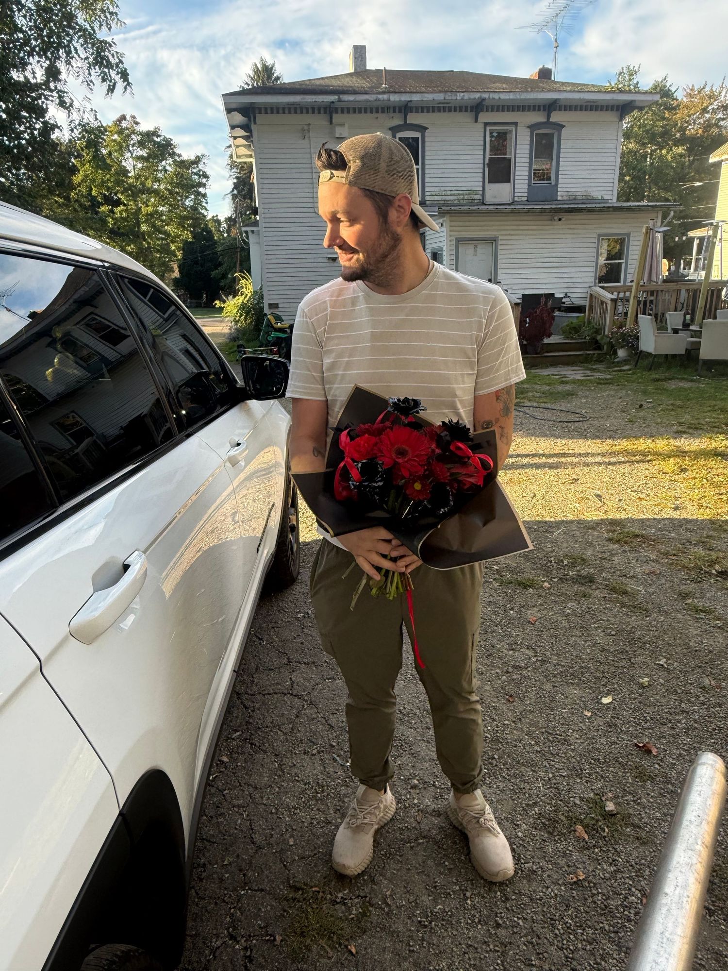 Man holding red roses bouquet in gravel driveway beside white SUV with house in background on sunny day.