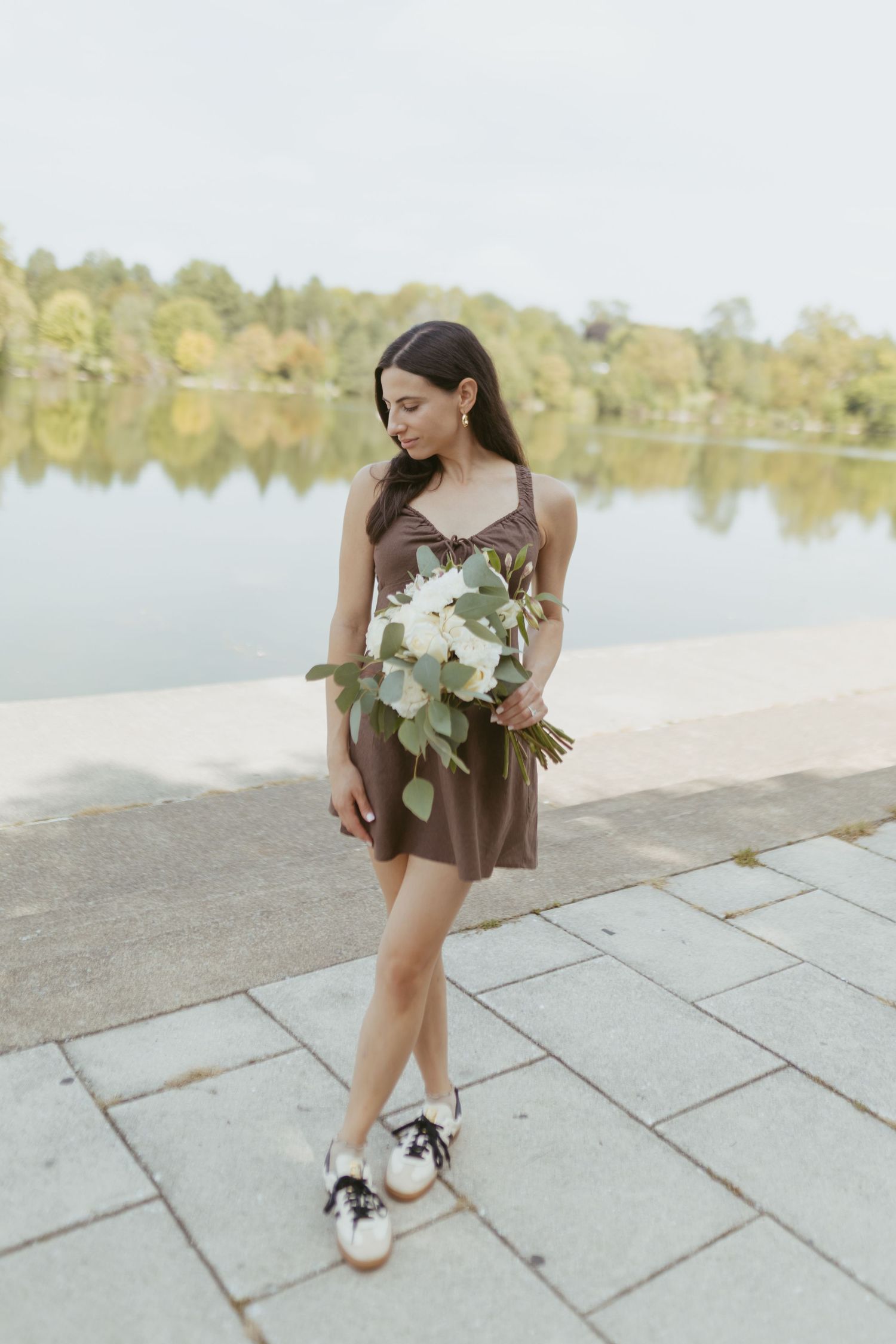 Young woman in brown mini dress holding white flower bouquet, standing by a serene lake in a park.