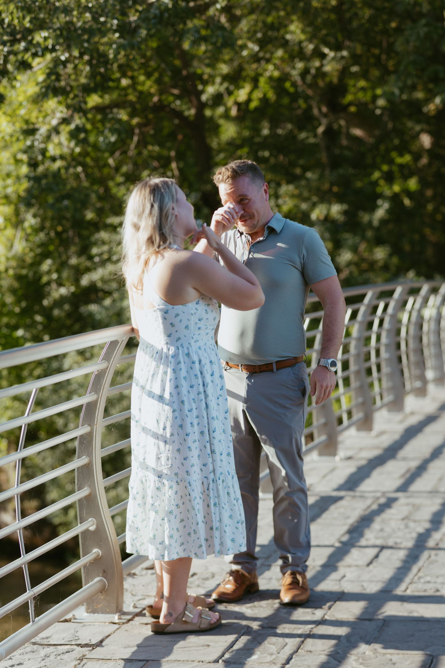 A couple shares a romantic moment on a modern bridge surrounded by lush green trees on a sunny day.