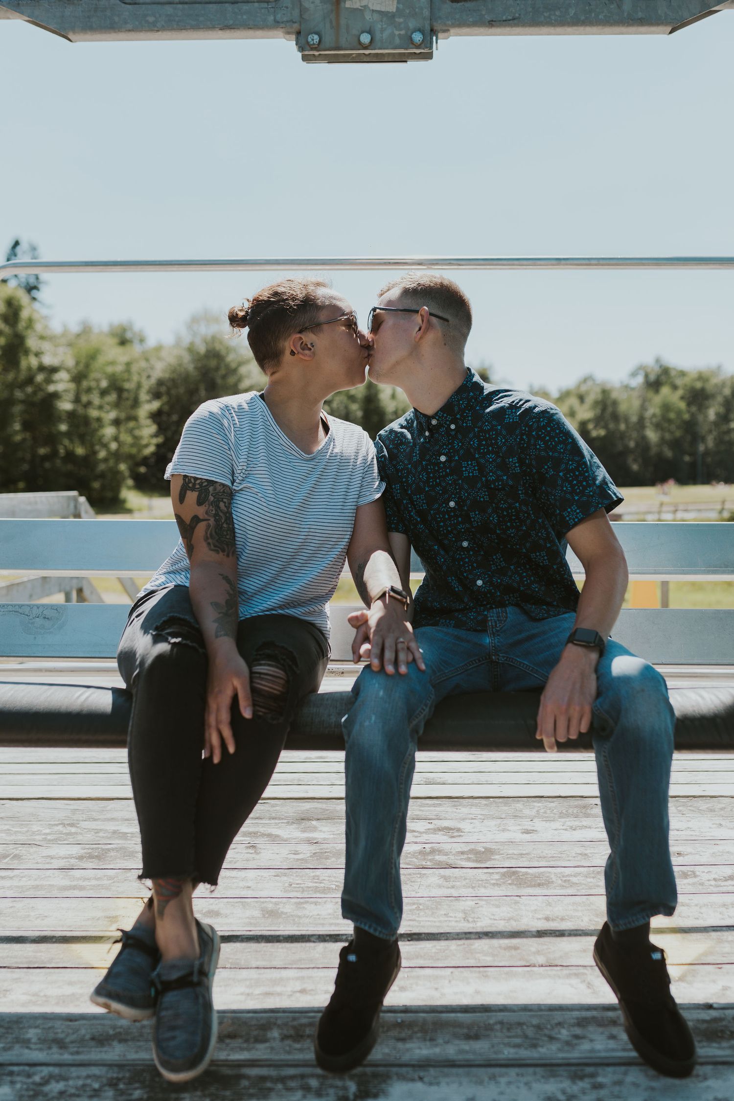 Couple sharing a kiss while sitting on bleachers outdoors on a sunny day, one with tattoos wearing a striped tee.
