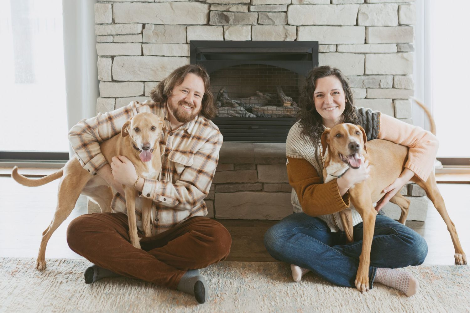 Couple sitting cross-legged by stone fireplace, each holding a golden dog, smiling warmly in cozy home setting.