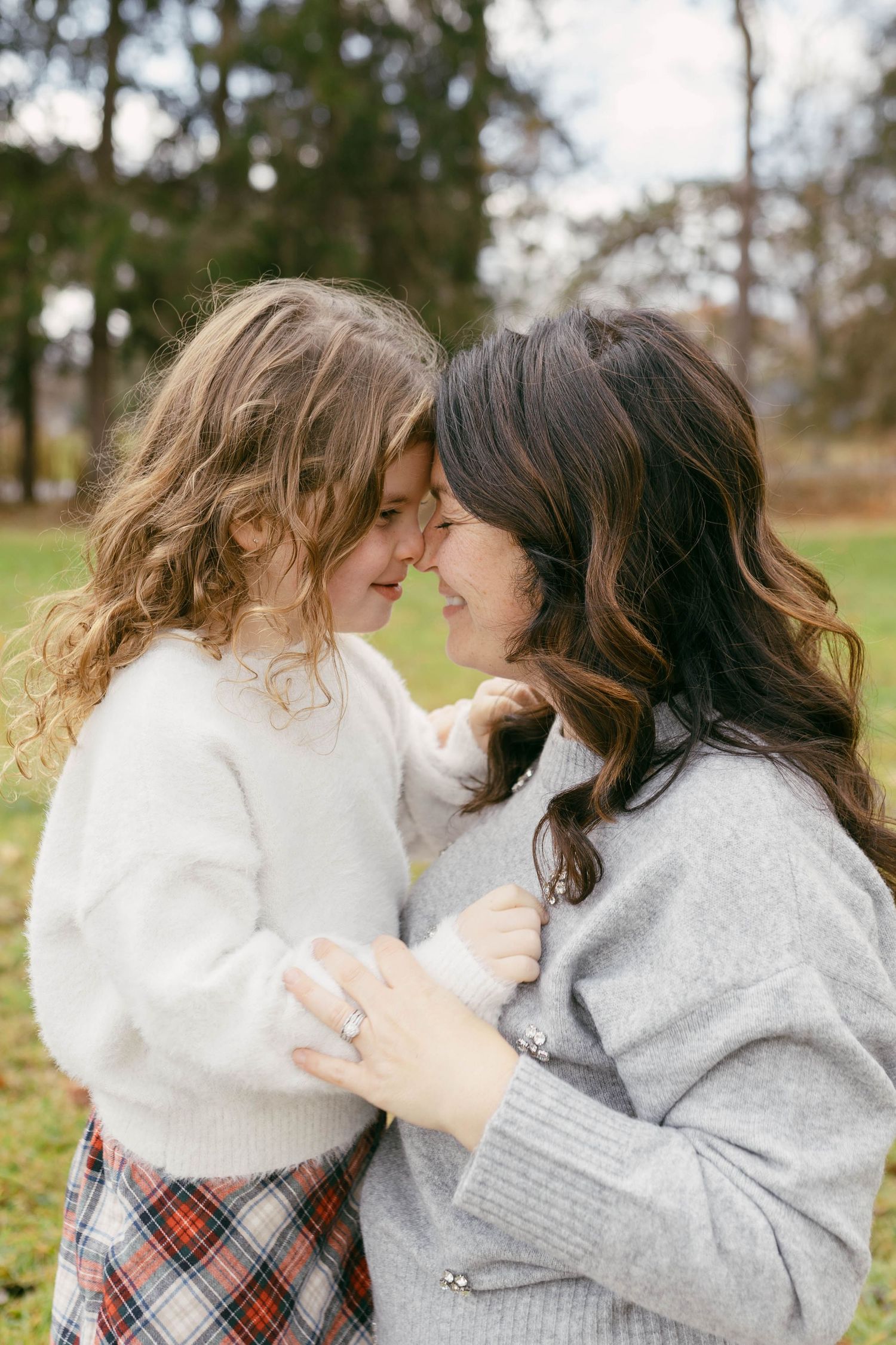 A mother and young daughter touch foreheads and smile outdoors, wearing cozy sweaters in a park setting.