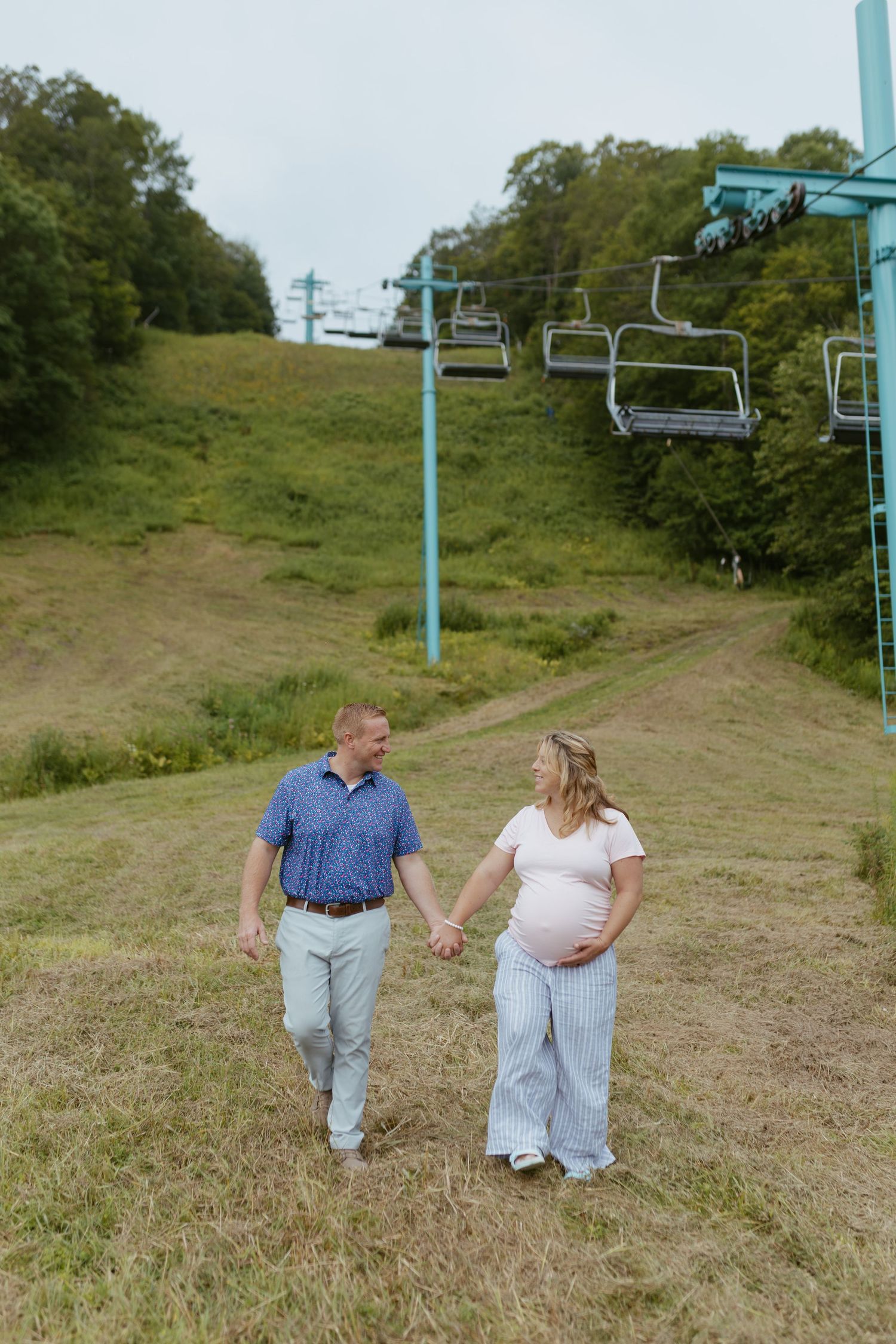 Pregnant couple holding hands walking on grassy hillside near ski lift chairs on overcast day.