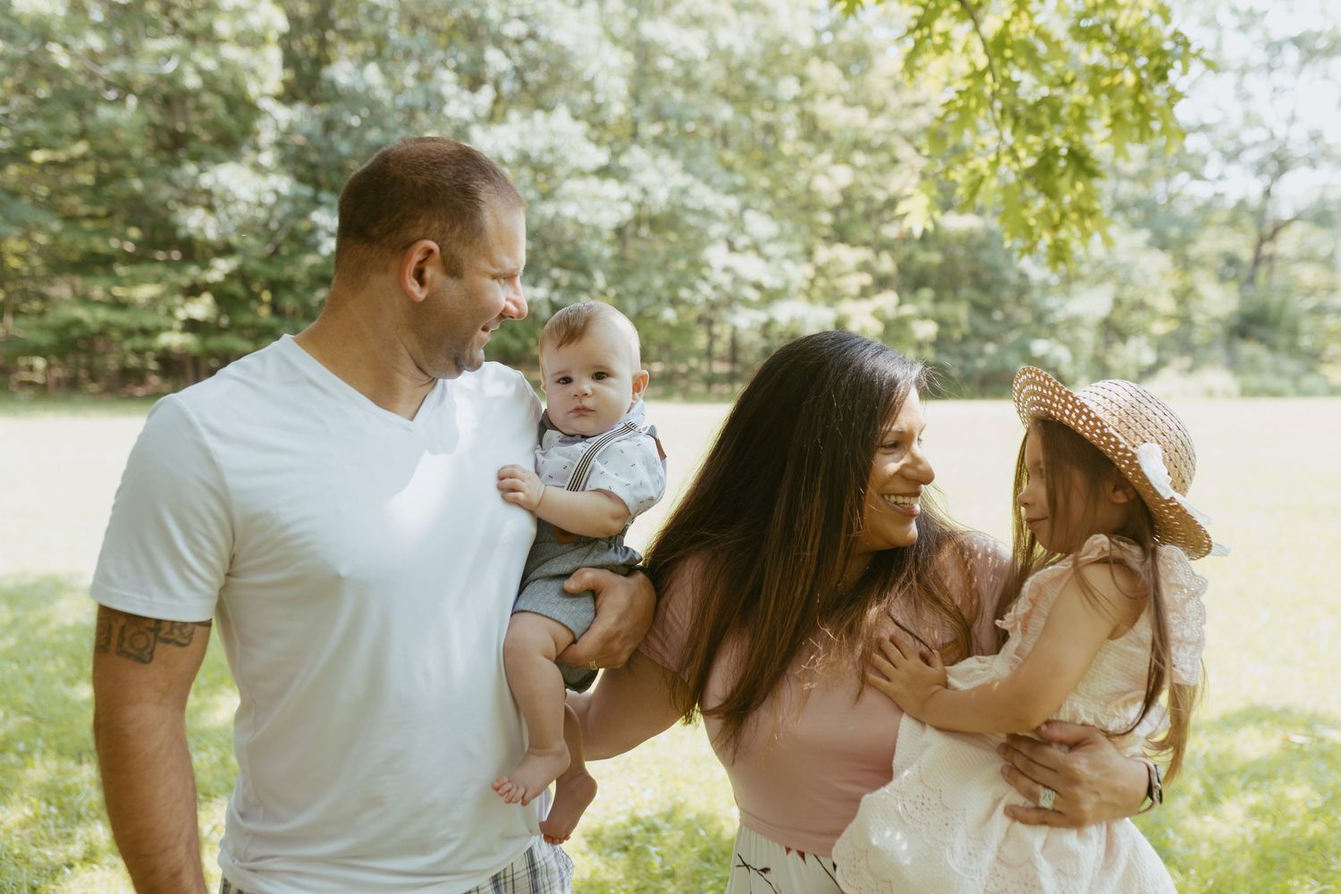 Happy family of four enjoying a sunny day outdoors, parents holding baby and toddler girl in a lush green park.