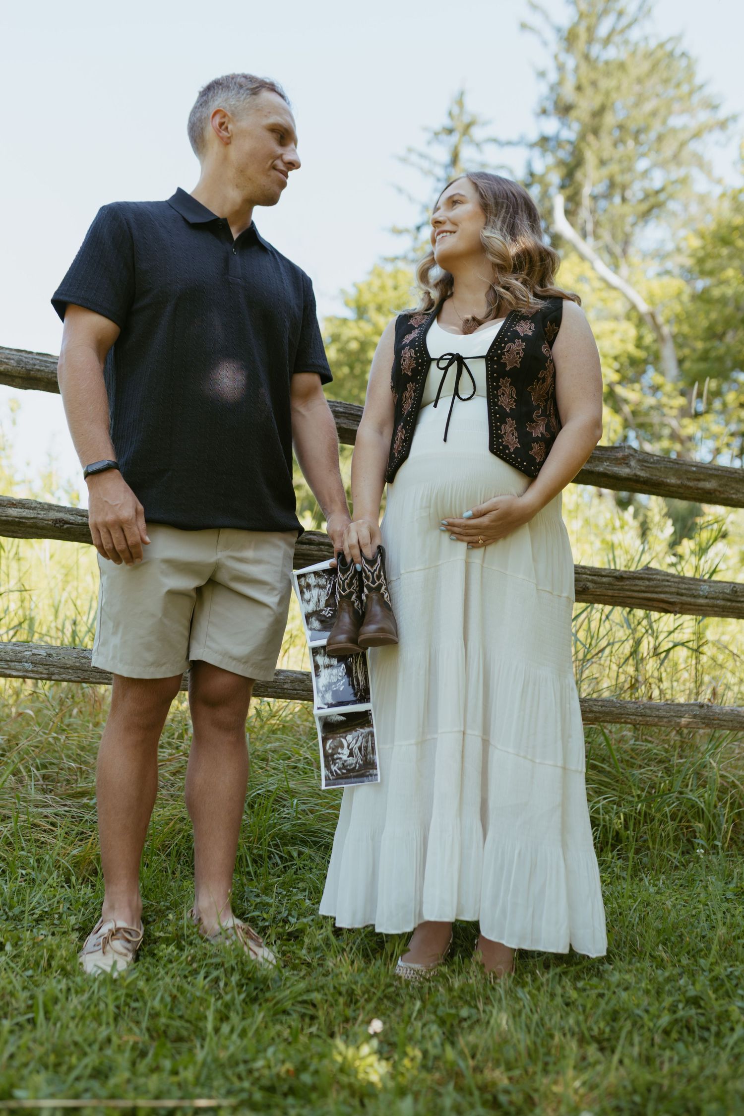 Pregnant couple holding ultrasound photos by rustic fence outdoors.
