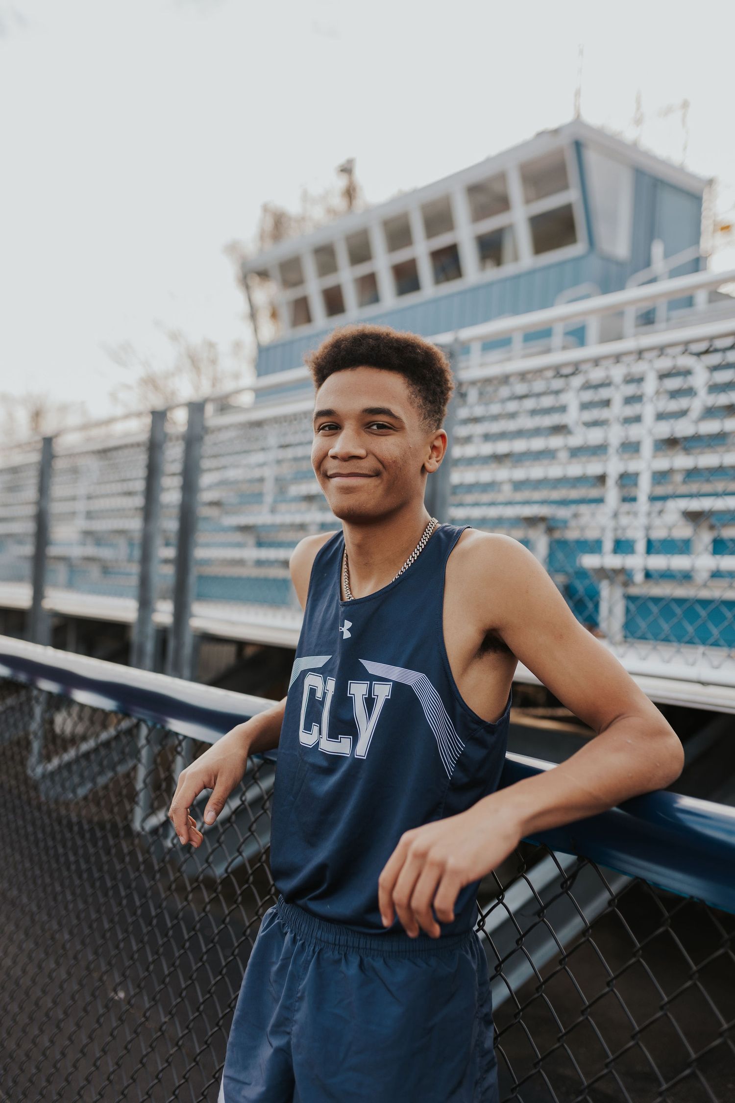 Young male athlete in navy CLV tank top smiling at a stadium track, leaning on a fence.