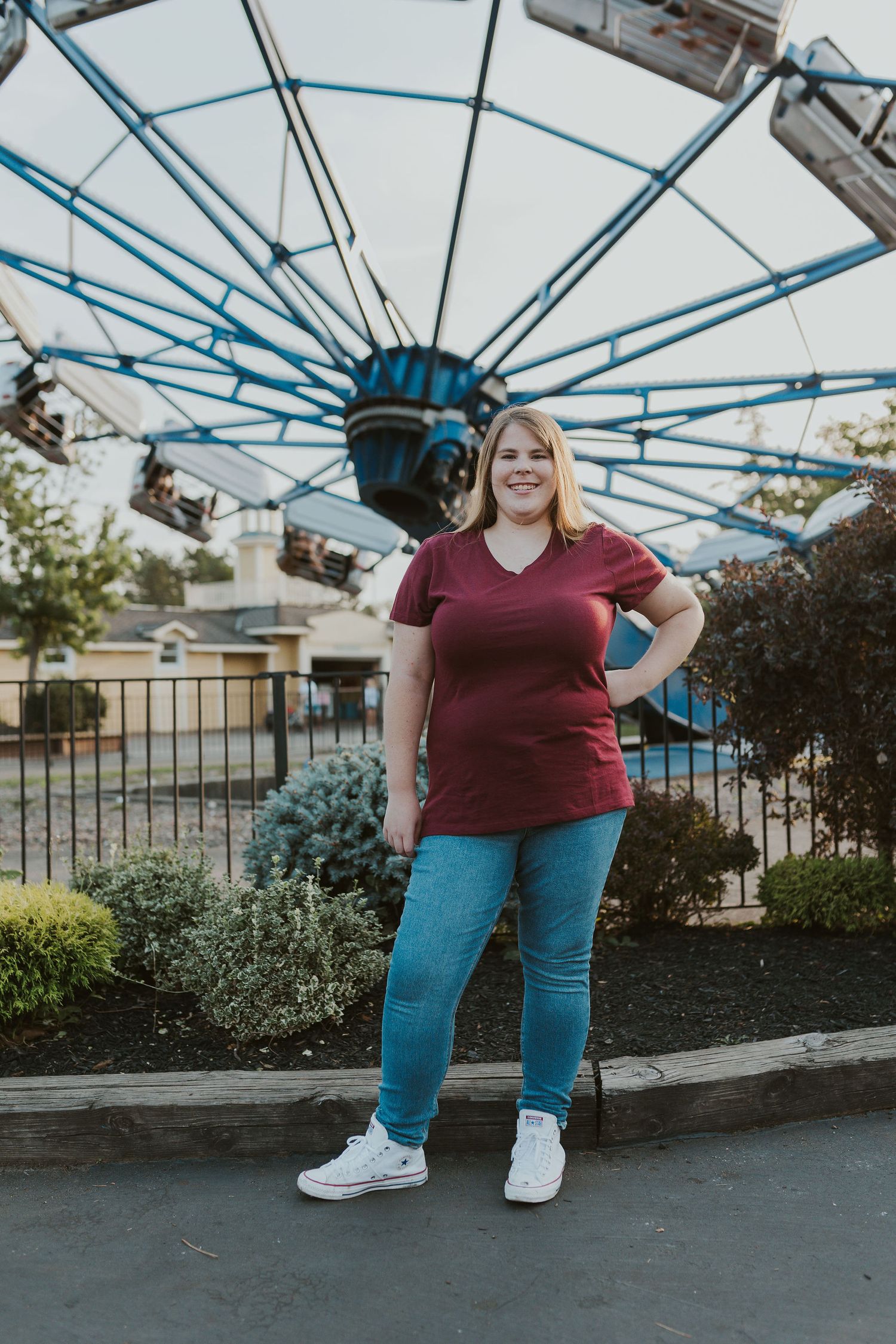 Woman in maroon shirt and jeans posing in front of a colorful ferris wheel at an amusement park.