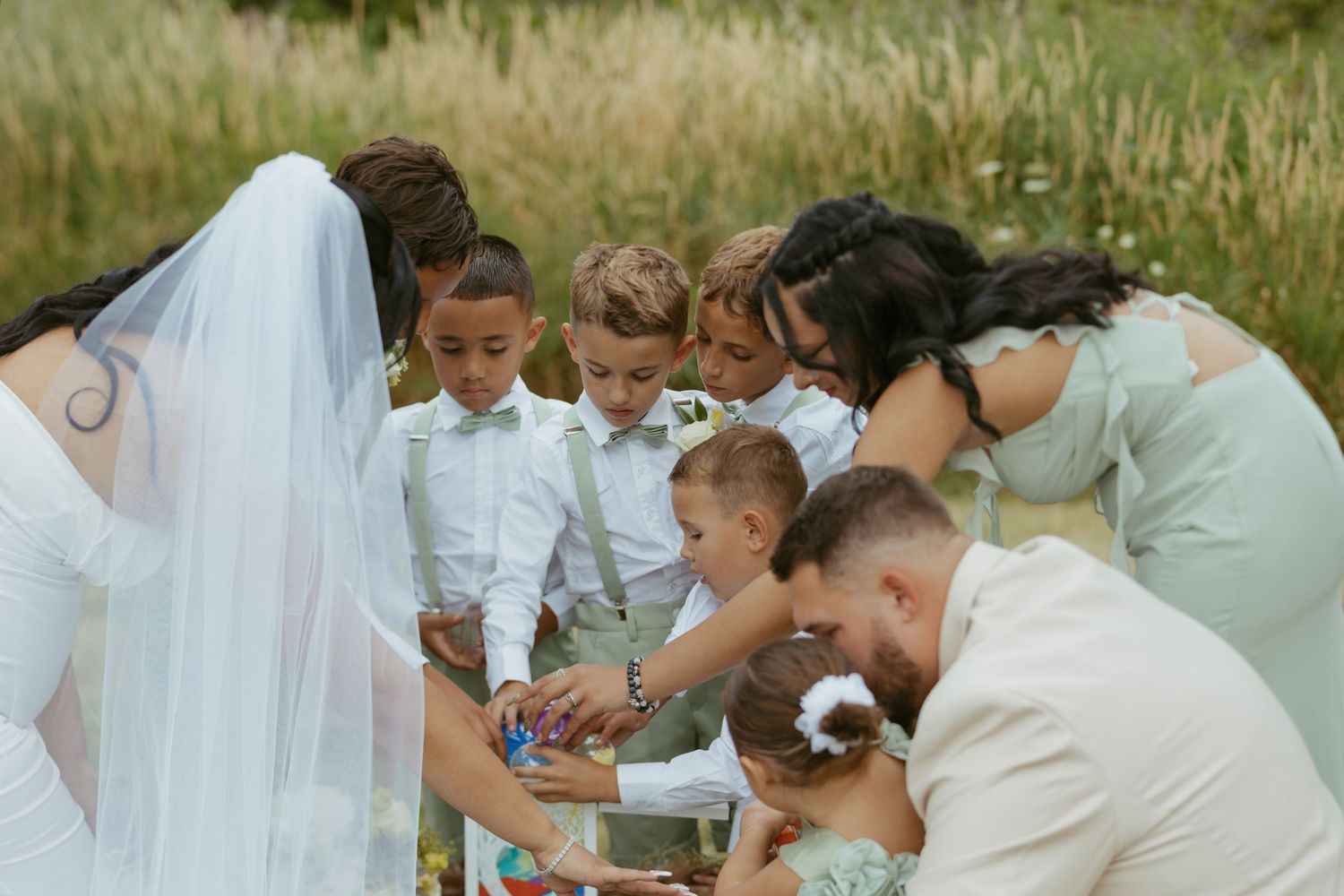 Wedding party and children gather outdoors for a unity ceremony in a sunlit field, bride in white veil with bridesmaids.