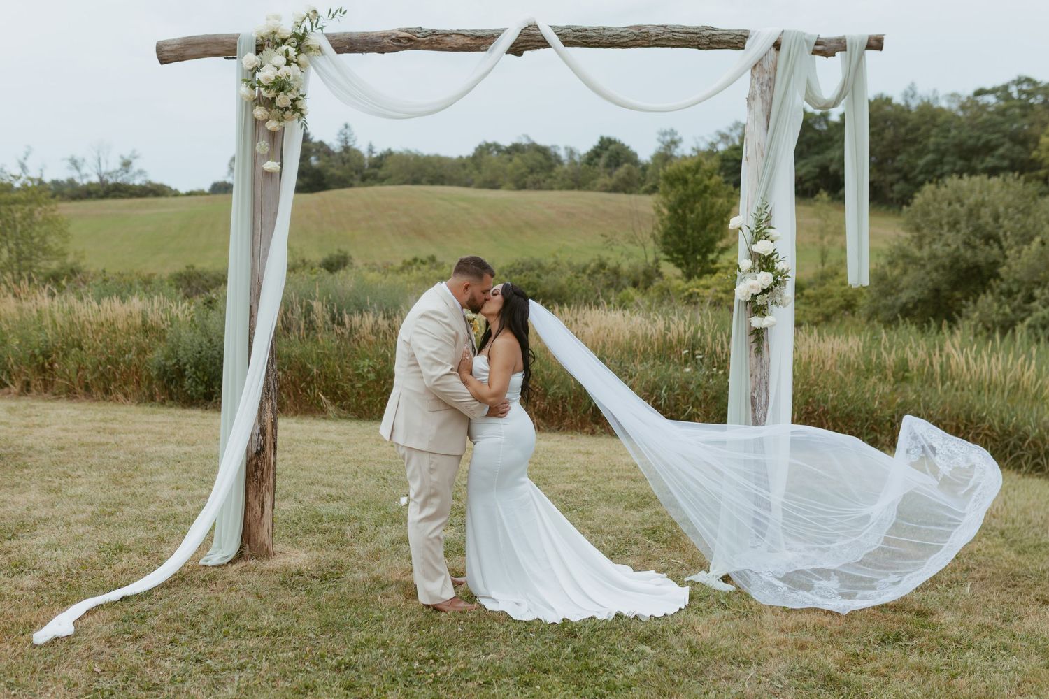 Bride and groom kissing under rustic wooden arch with white draping and floral accents in scenic outdoor wedding ceremony.