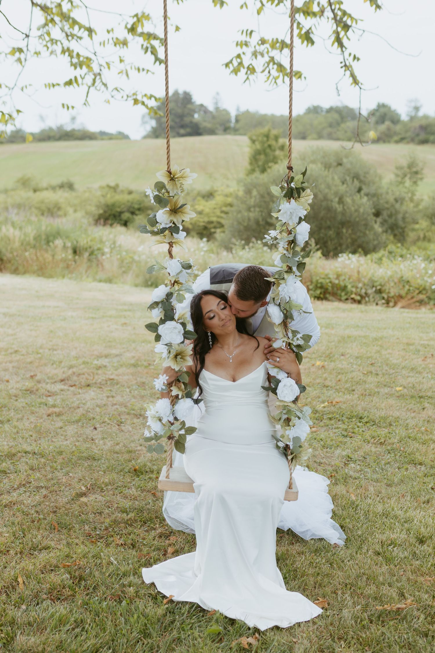 Bride in white gown sits on floral swing as groom kisses her cheek outdoors at a romantic garden wedding.