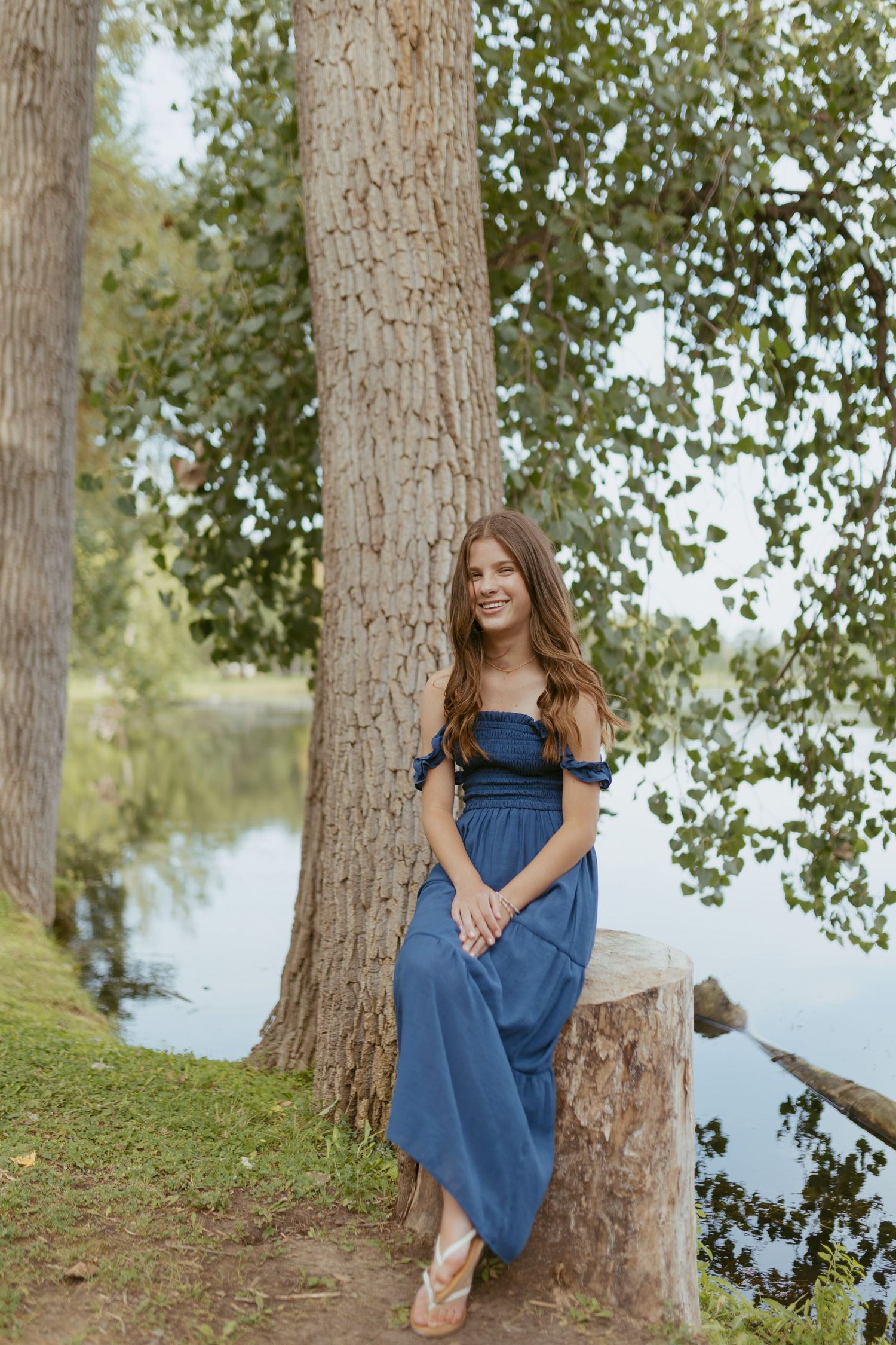Young woman in blue off-shoulder jumpsuit leaning against a tree by a serene lake in a lush park.