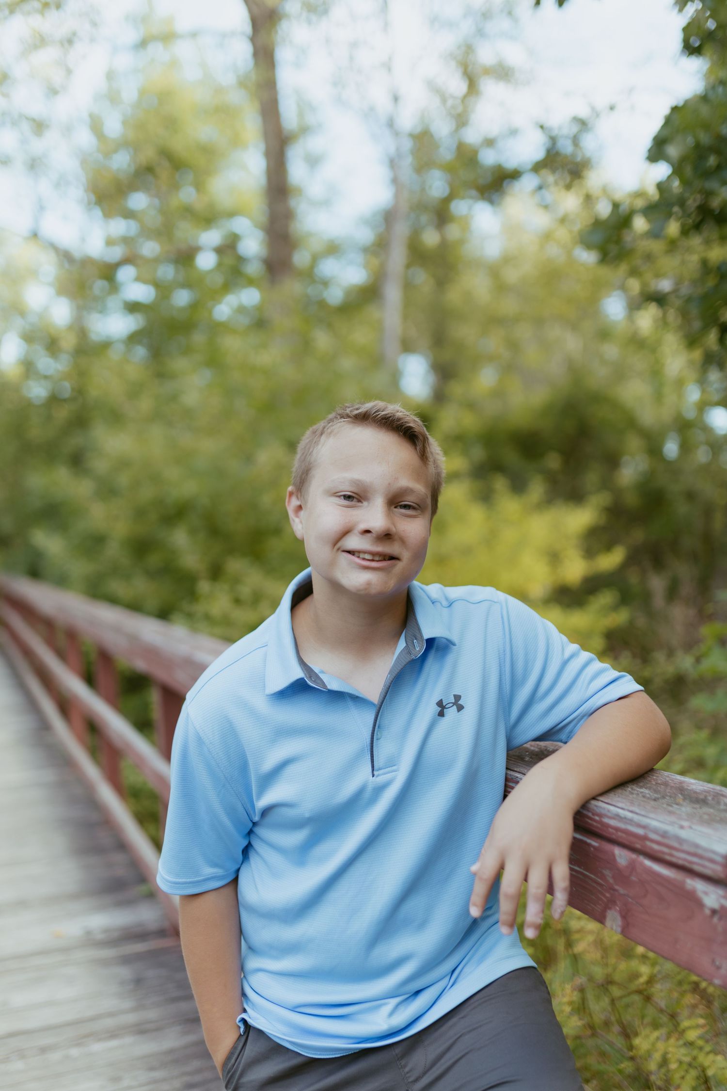 A smiling teenage boy in a light blue Under Armour polo leans on a wooden bridge railing surrounded by lush green trees.