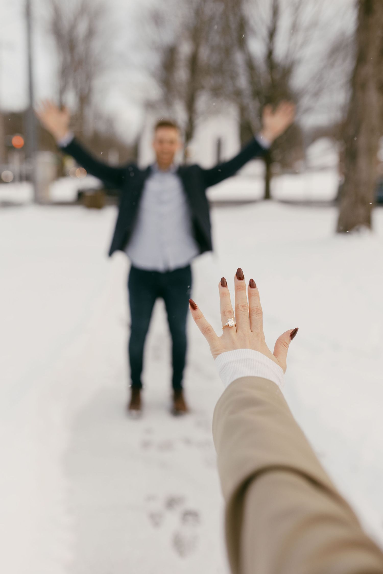 Woman showing off engagement ring in snowy park with excited man celebrating in background.