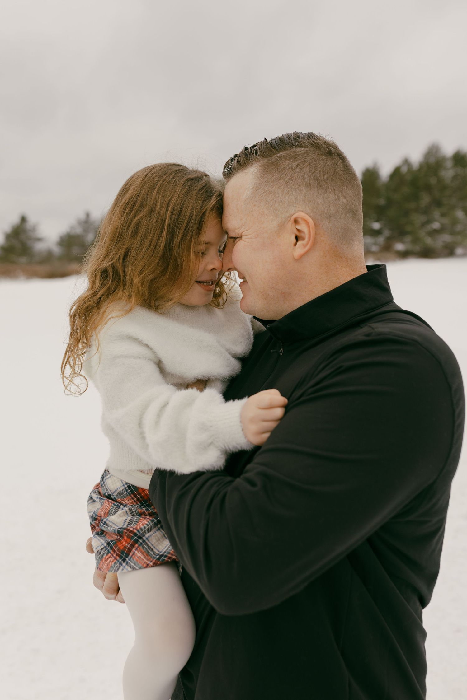 A father holds his young daughter close in a snowy winter landscape, both smiling and touching foreheads affectionately.