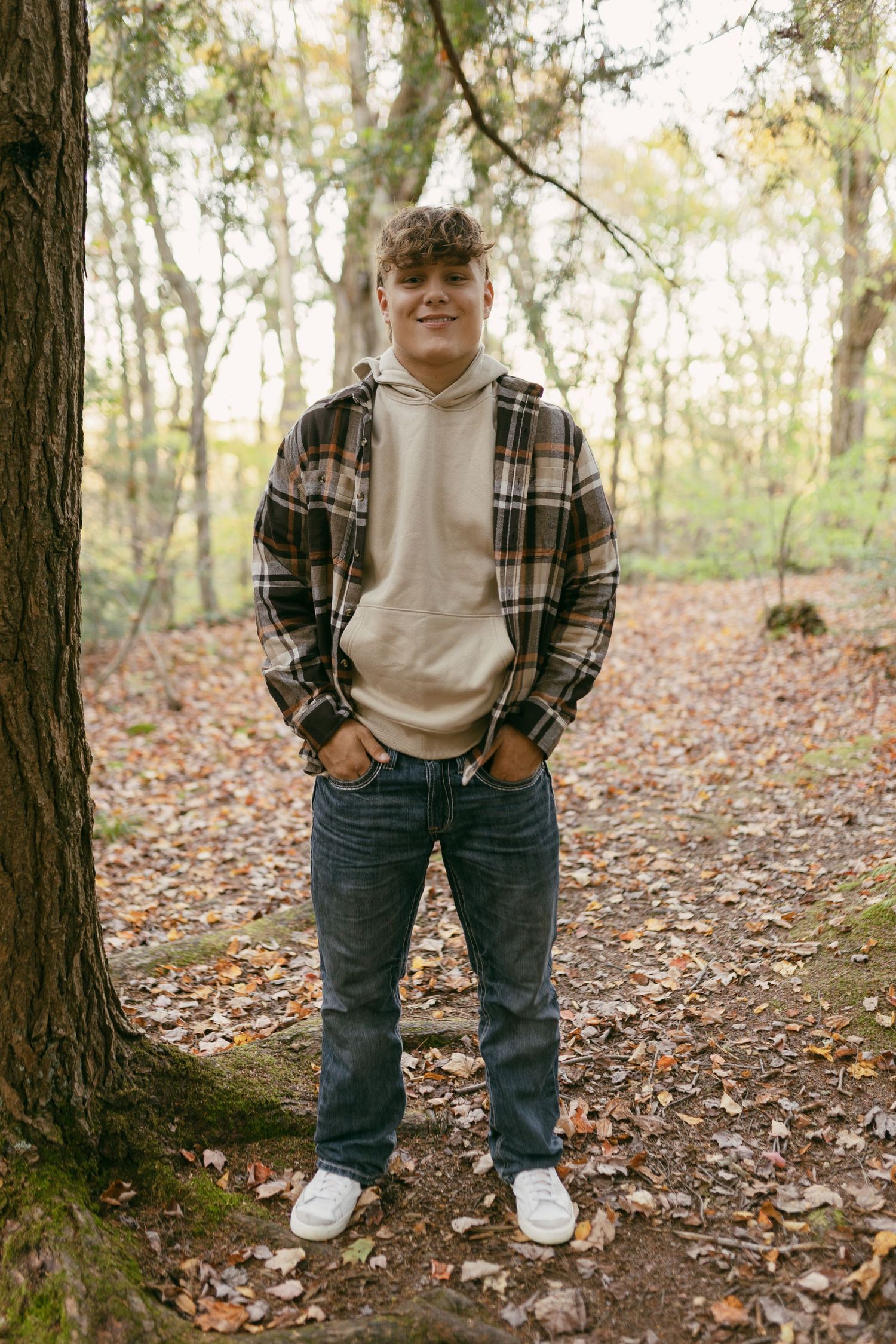 Teen boy in plaid shirt and hoodie standing in autumn forest with fallen leaves, hands in pockets.