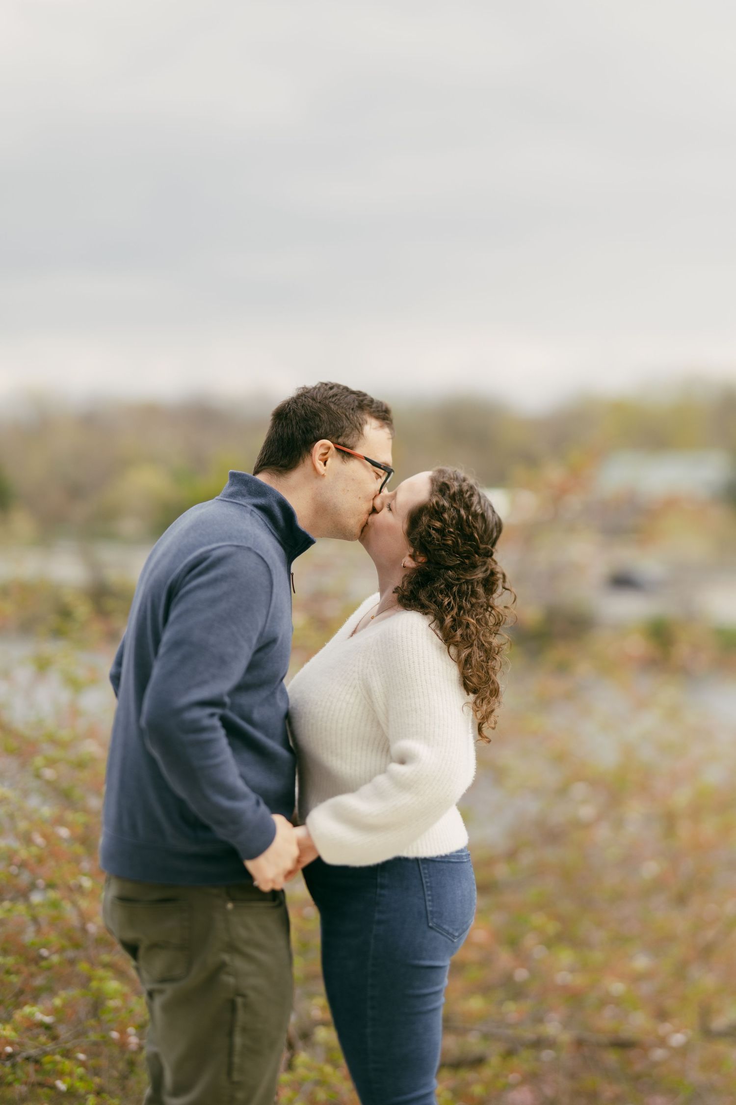 Couple sharing a romantic kiss outdoors in autumn, man in blue sweater and woman in white sweater holding hands.