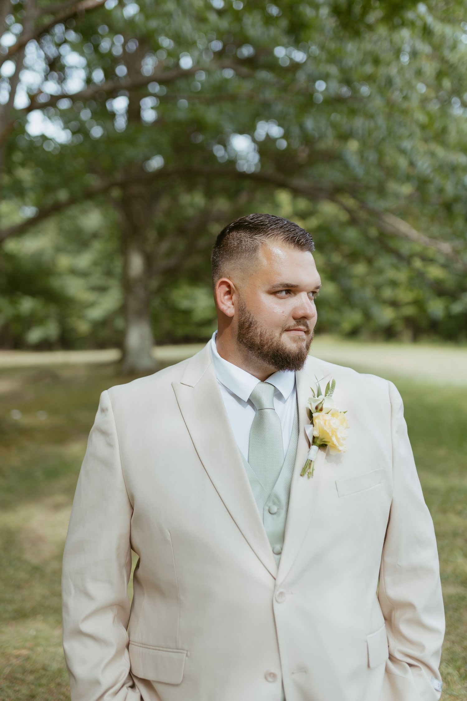 Groom in a cream suit with mint tie and boutonniere poses outdoors in a lush green park setting.