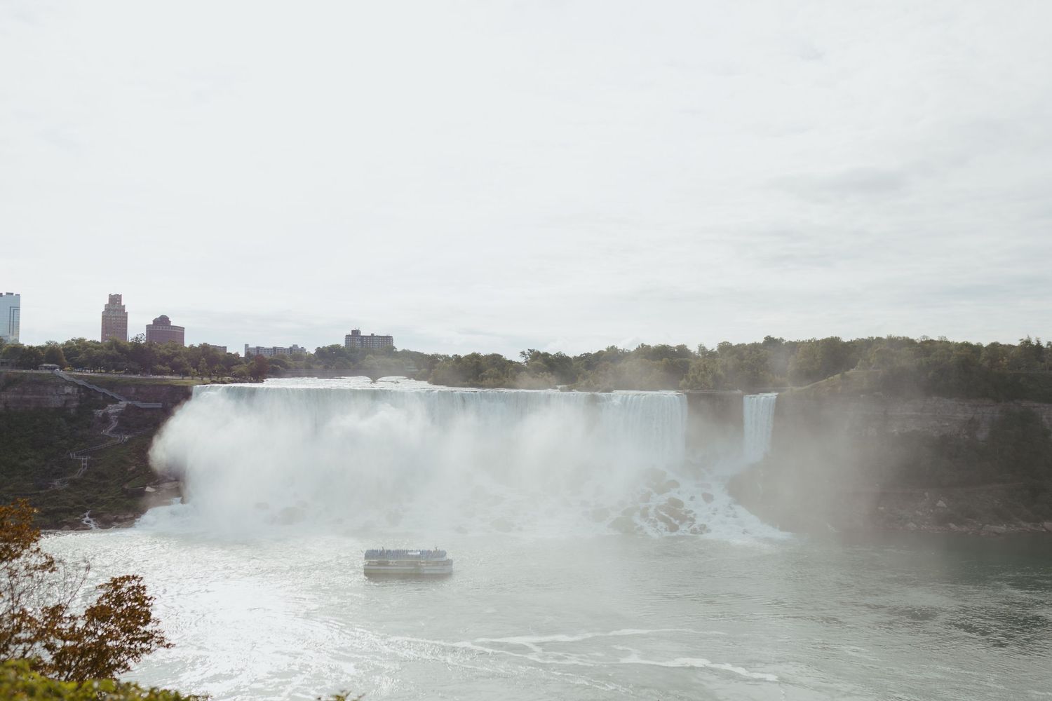 Misty Niagara Falls with powerful cascading water, surrounding trees, and city skyline in the background on an overcast day.