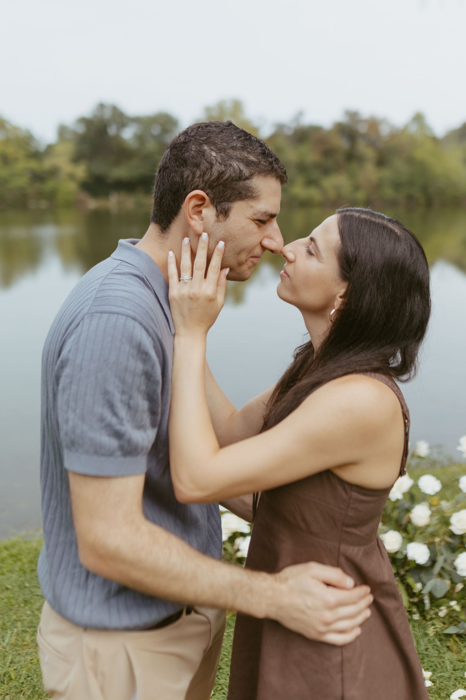 Couple sharing an intimate moment by a lake, woman showing engagement ring while embracing her partner.