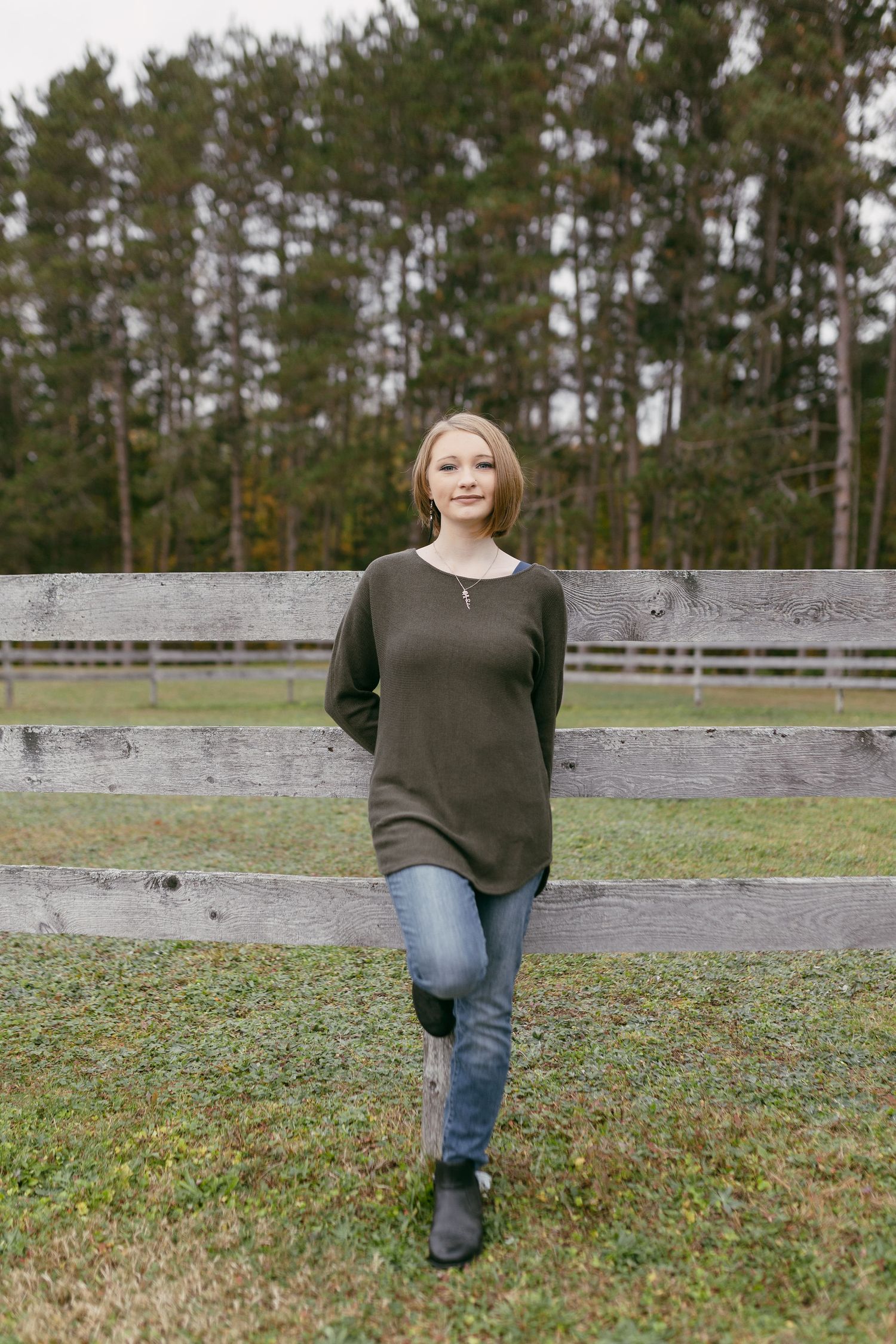 A person in a dark sweater and jeans poses in front of a stone wall and grassy field.