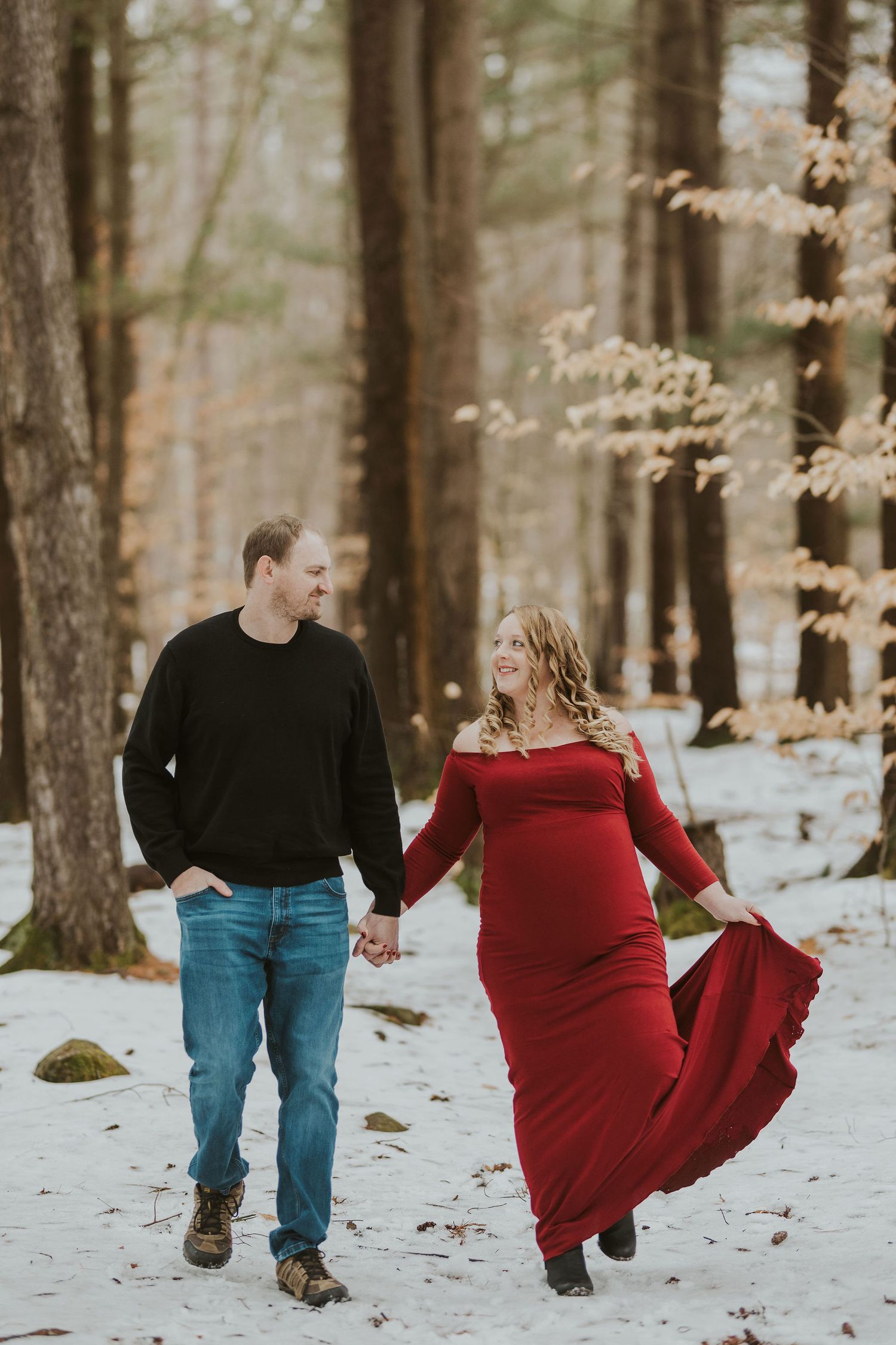 Couple walking hand in hand through snowy forest with woman wearing flowing red dress.