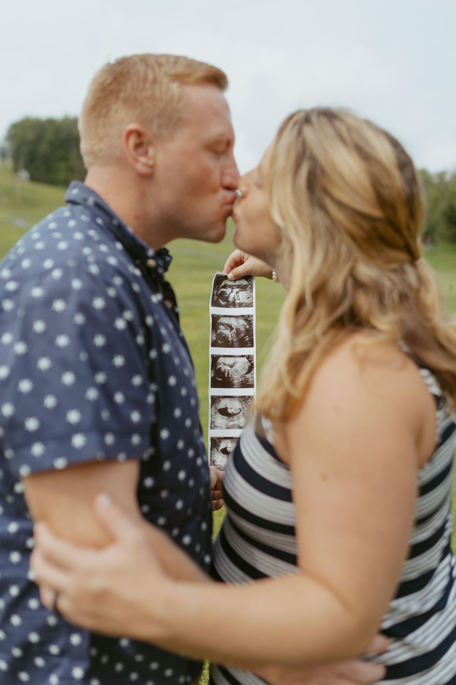 Couple kissing while holding ultrasound photos, pregnancy announcement in a green outdoor field setting.