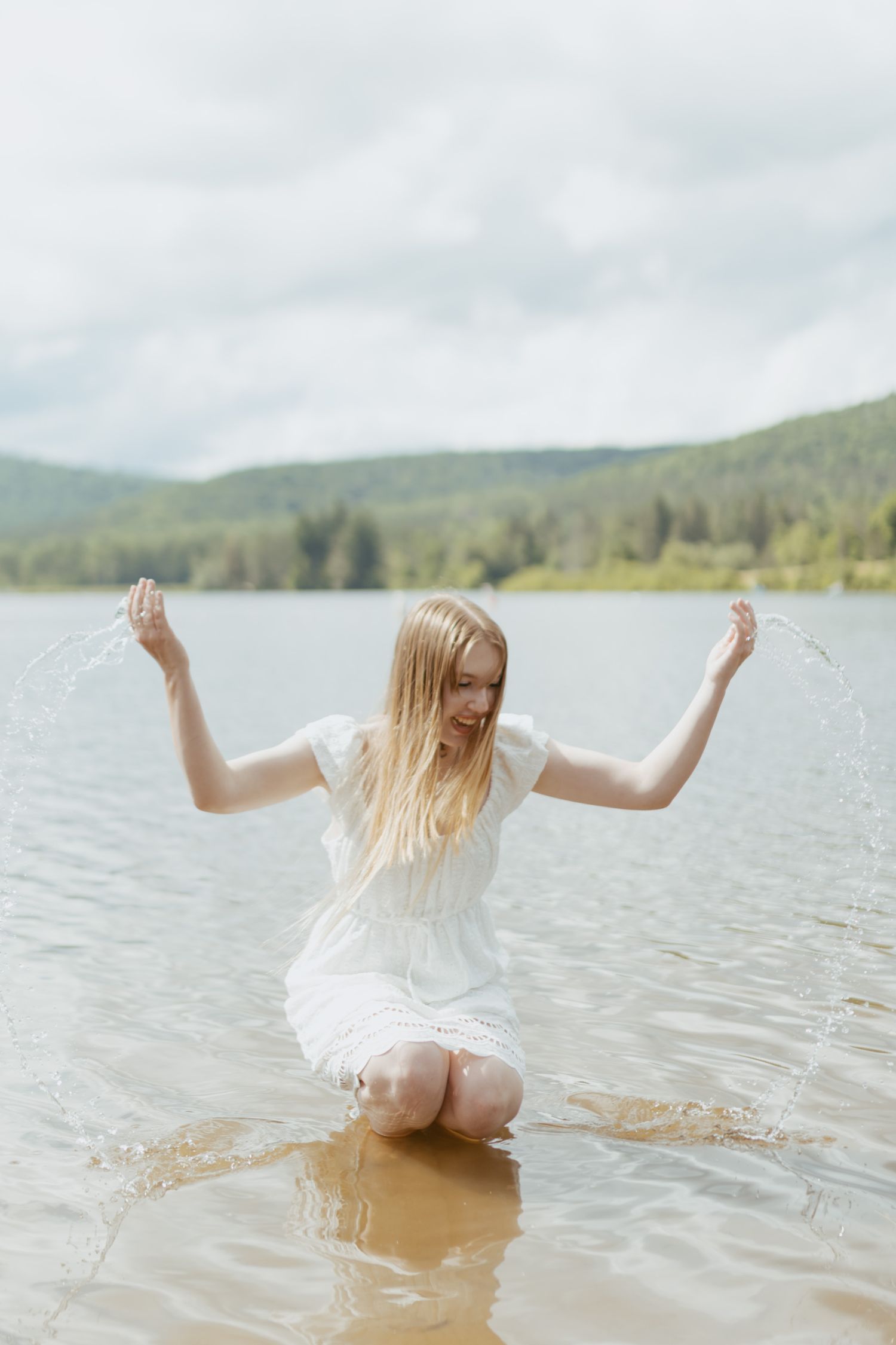 Young blonde woman in white dress kneeling in a lake, joyfully splashing water with mountains in background.