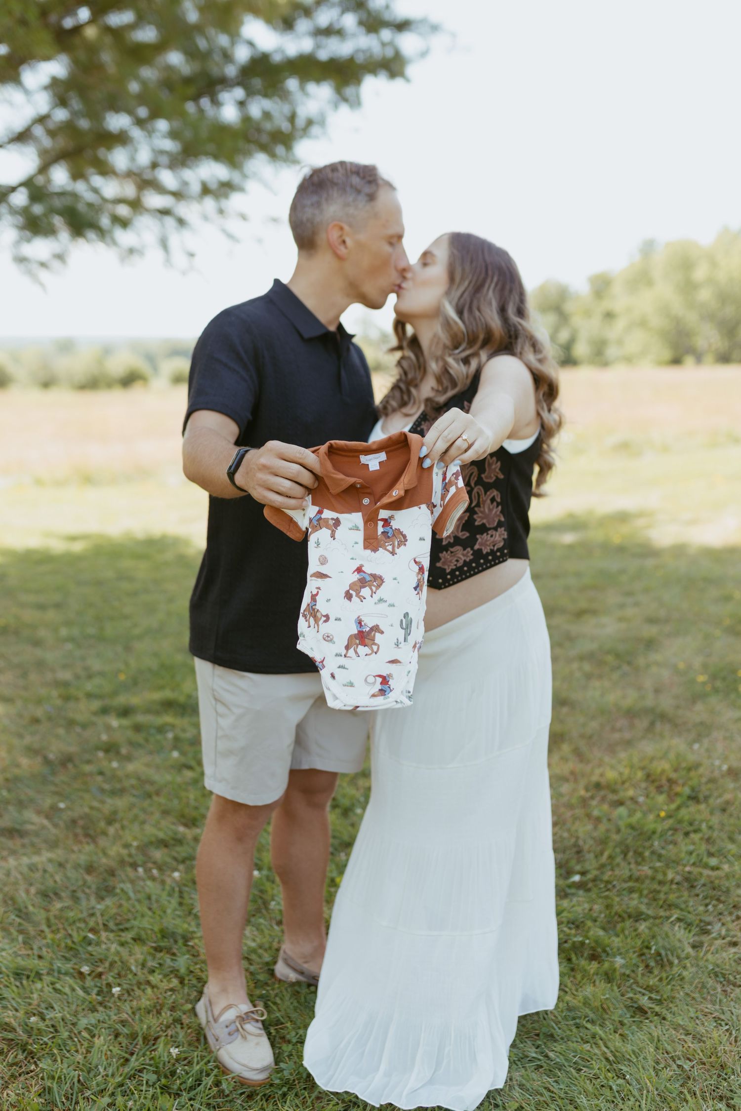 Couple kissing outdoors holding a baby onesie, announcing pregnancy in a sunny green field.