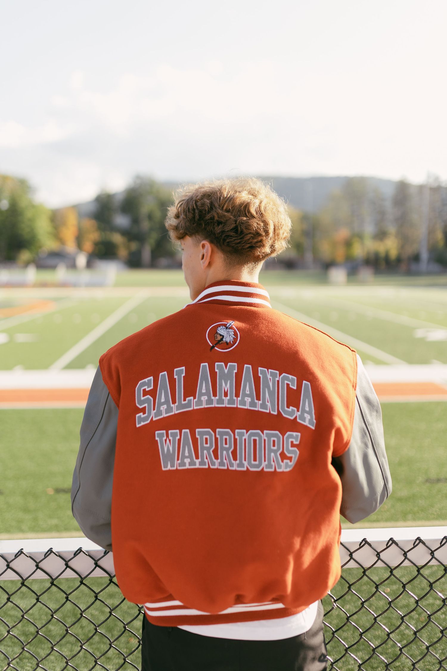 Student wearing an orange Salamanca Warriors varsity jacket stands overlooking a green football field.