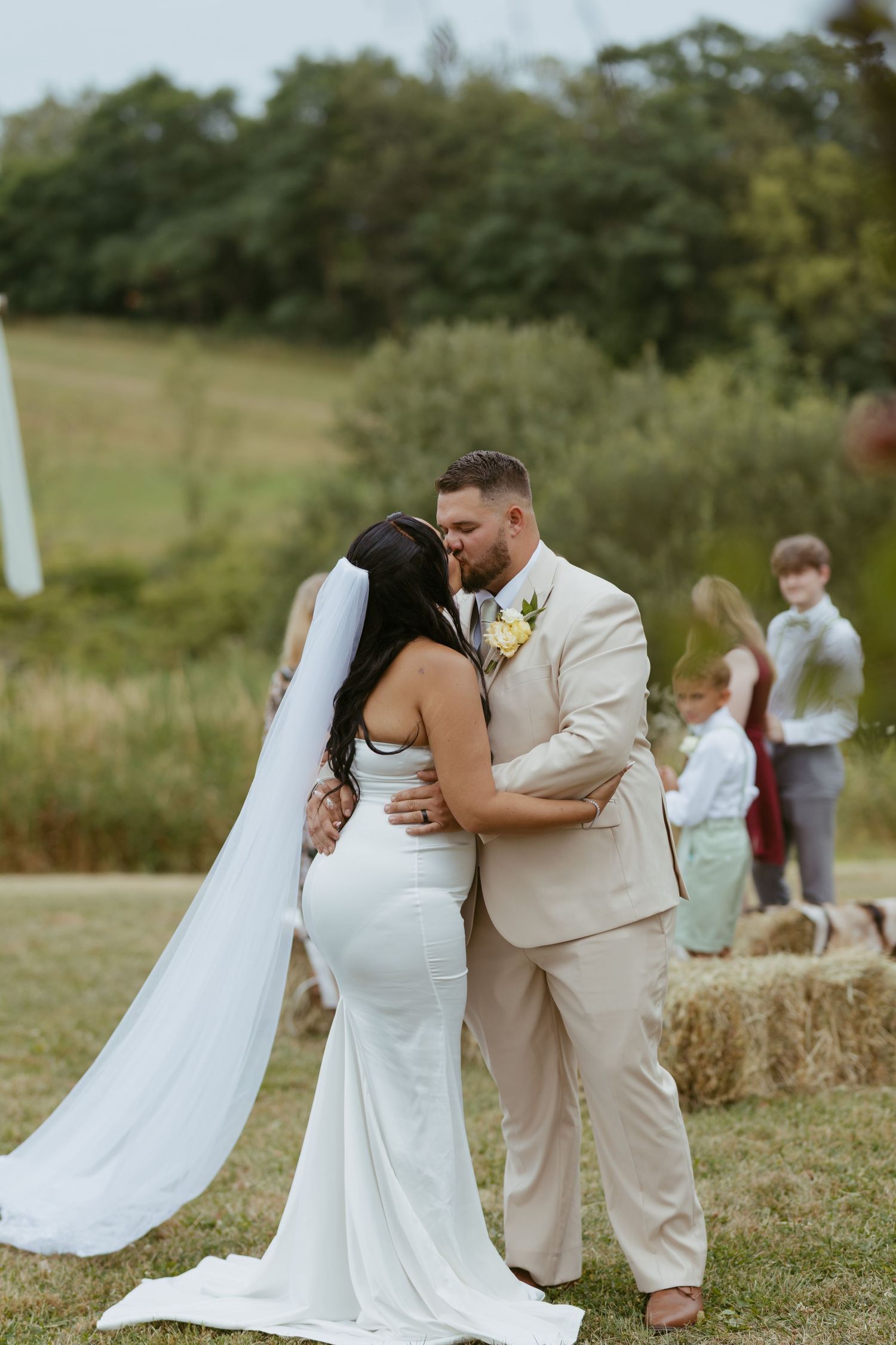 Bride and groom share first kiss at outdoor rustic wedding ceremony with hay bales and green hills backdrop.