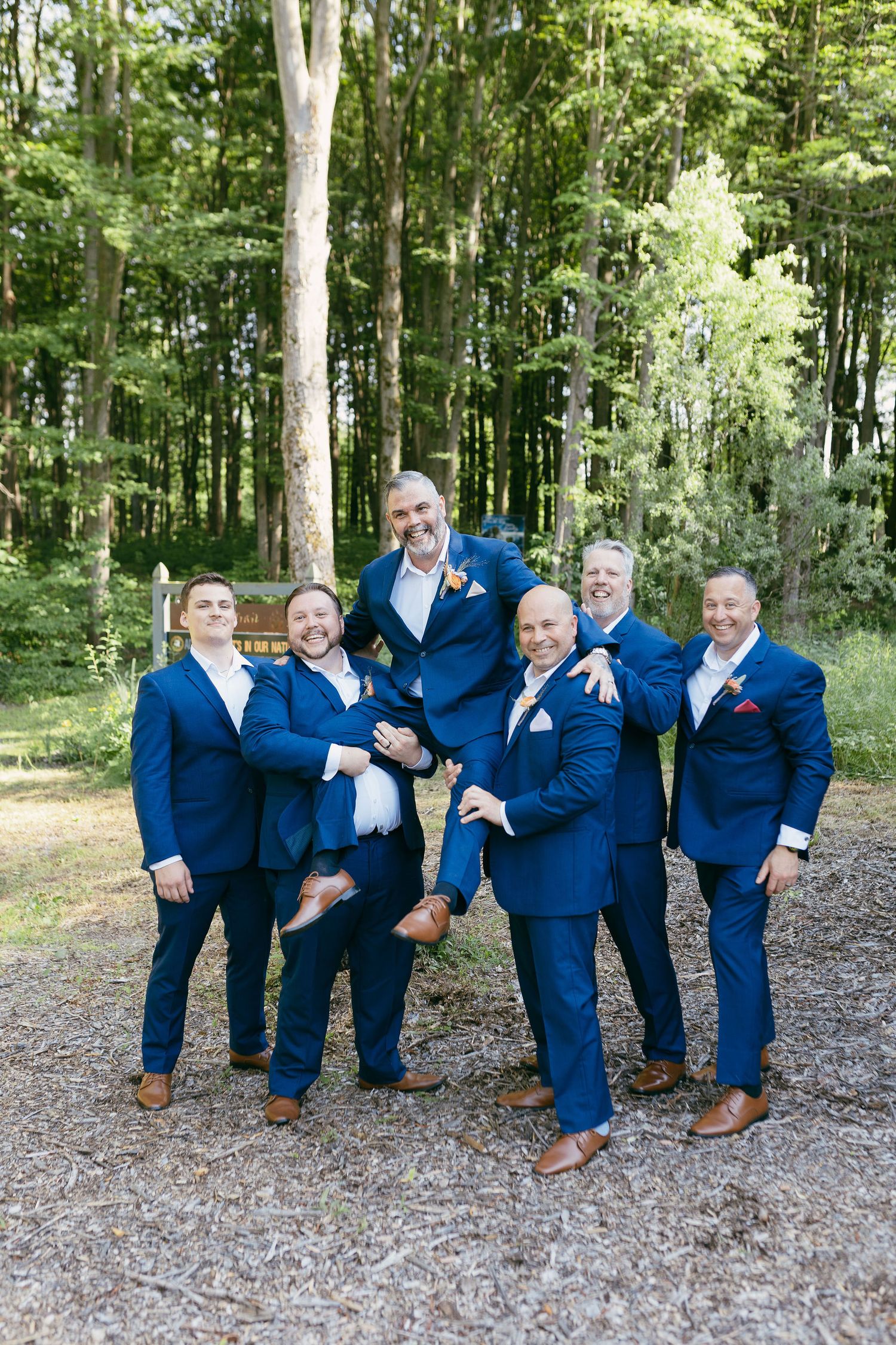 Groomsmen in navy suits strike playful poses together in a wooded outdoor setting.