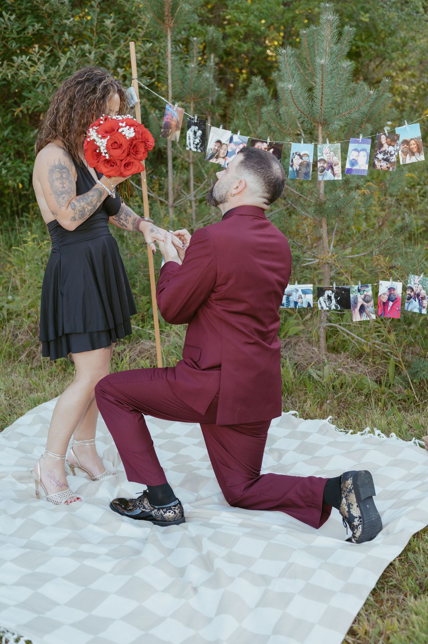 Man in burgundy suit proposes on one knee to woman in black dress holding red bouquet, outdoors with photo garland backdrop.
