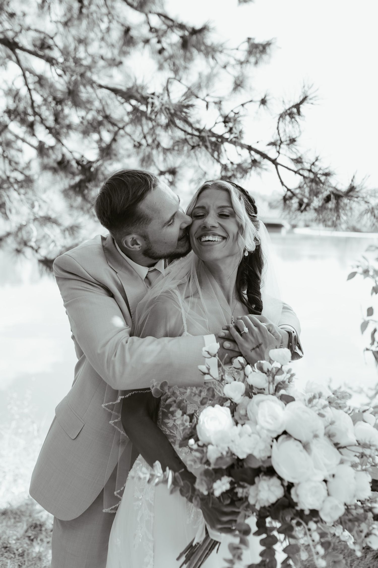 Black and white photo of a groom kissing bride on cheek as she laughs holding a large floral bouquet outdoors.
