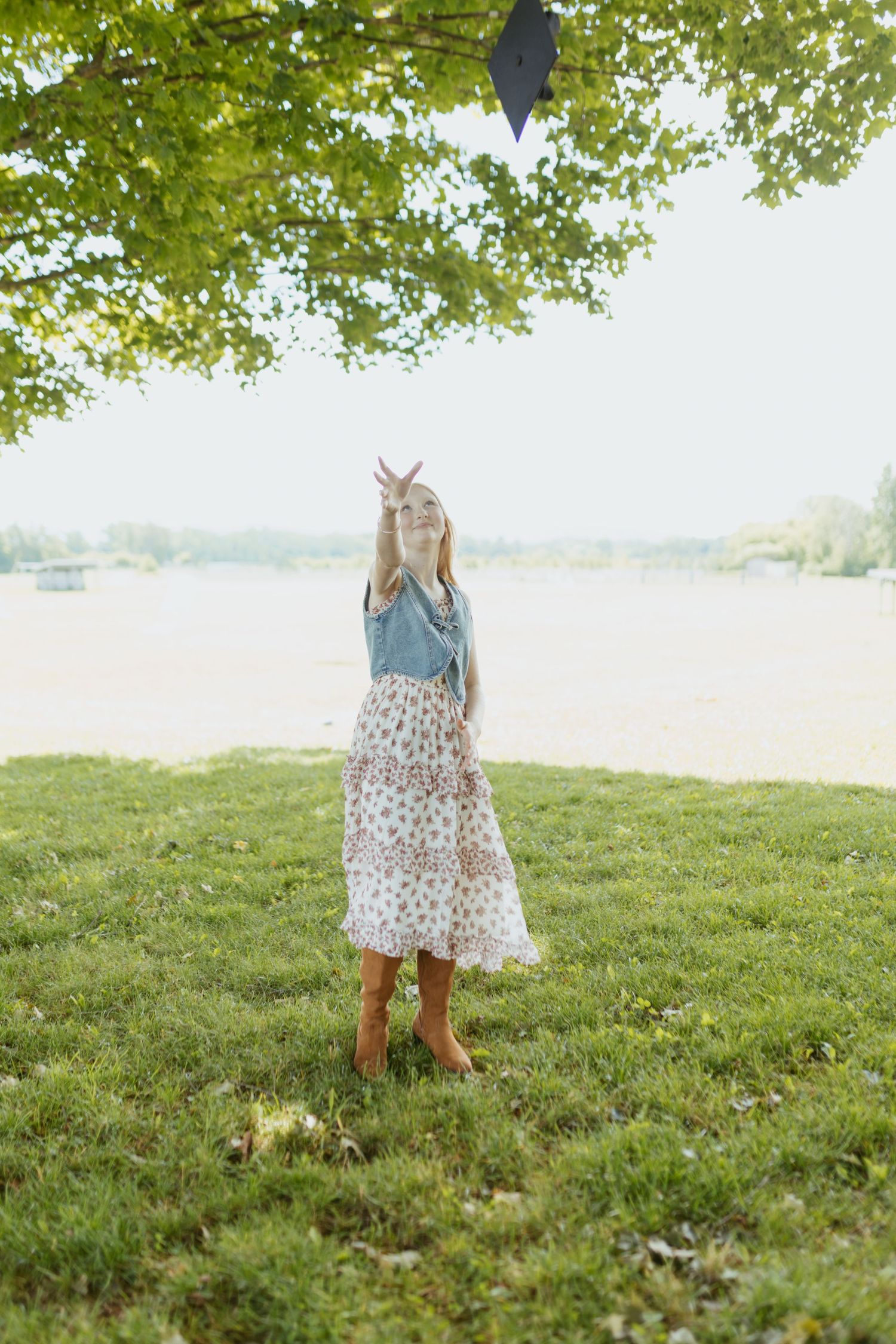 A young girl in a floral skirt and denim top tosses a graduation cap outdoors near a lake.