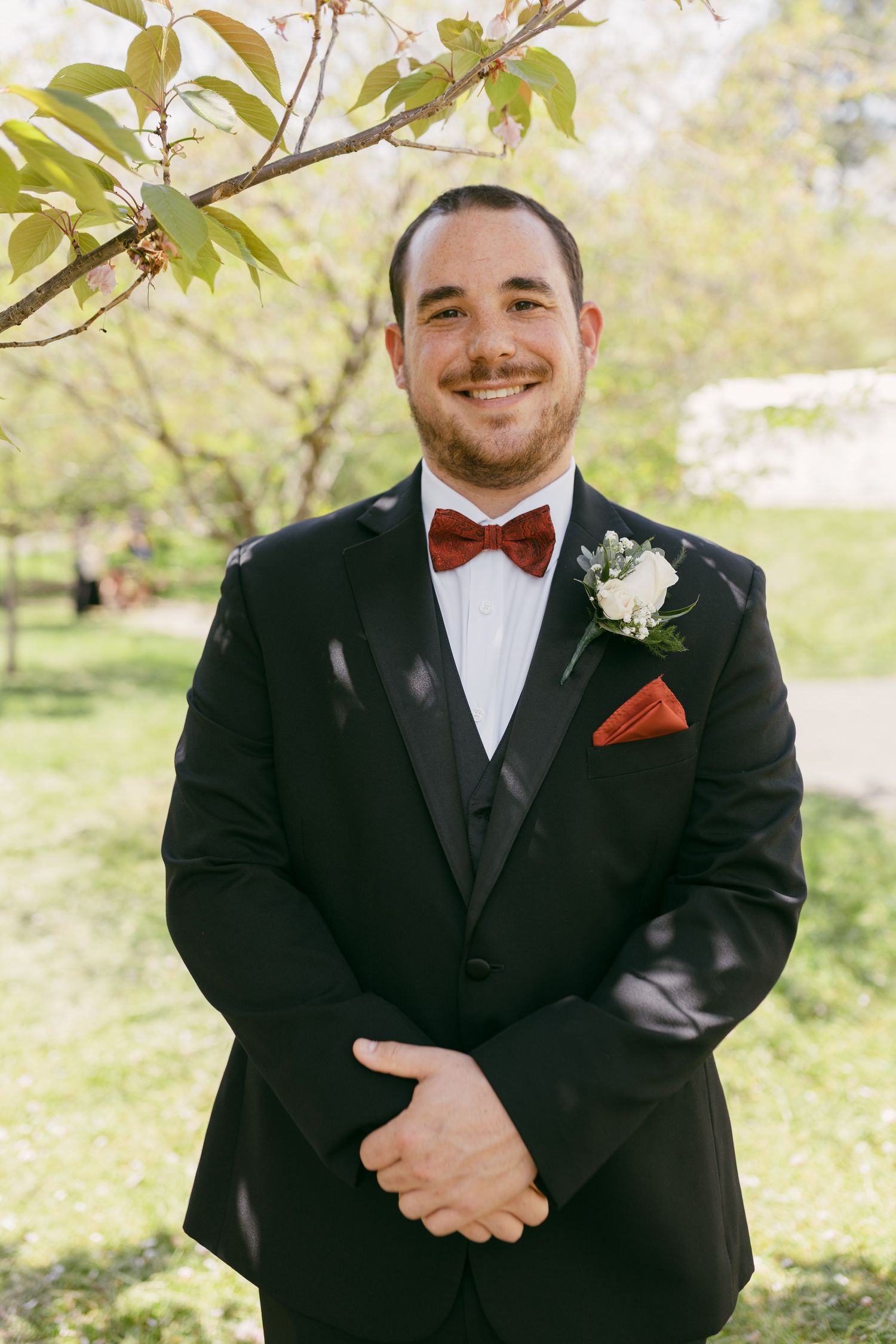 Smiling groom in black tuxedo with red bow tie and white boutonniere posing outdoors under a tree.