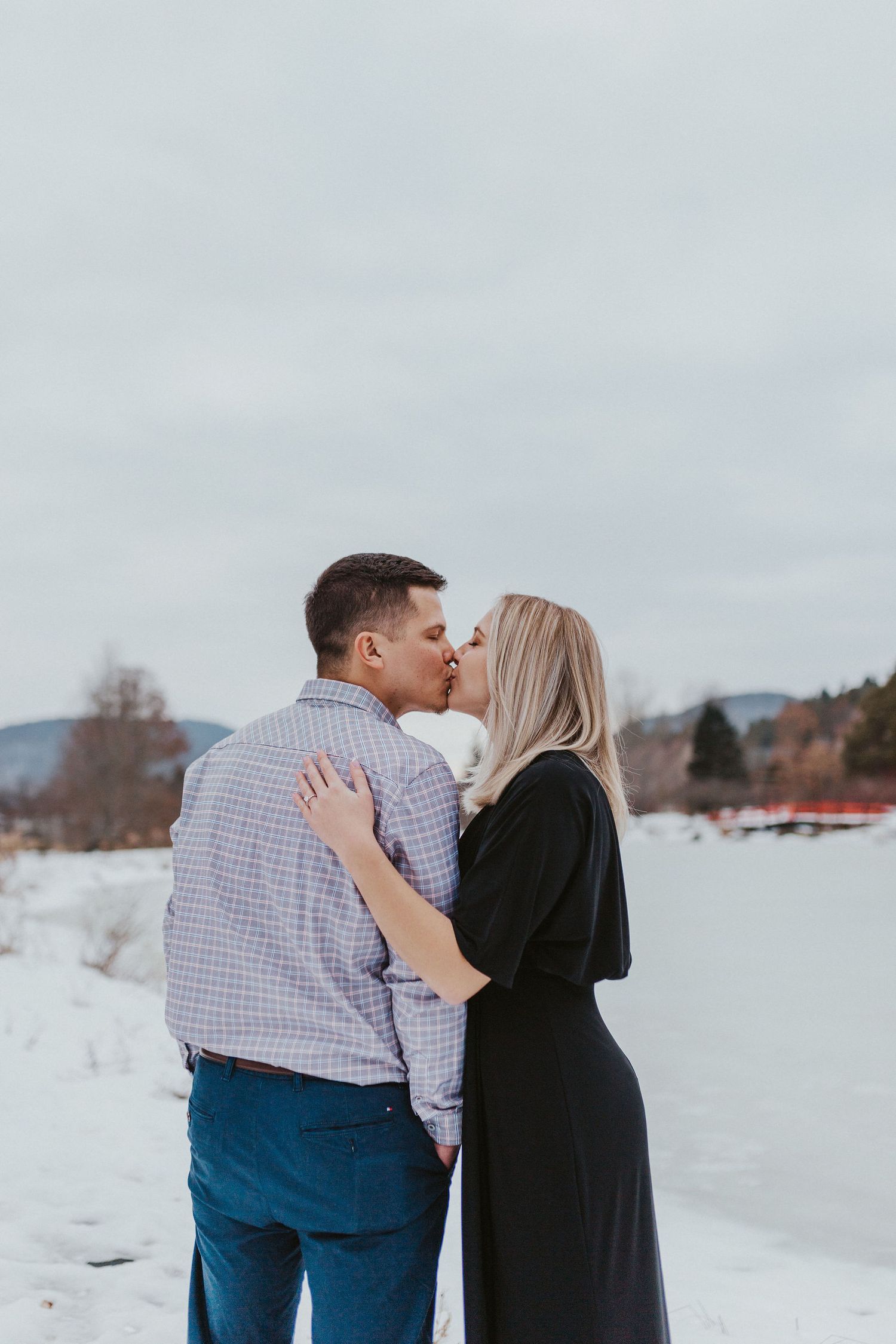 Couple sharing a romantic kiss in a snowy winter landscape, woman in black dress, man in plaid shirt.
