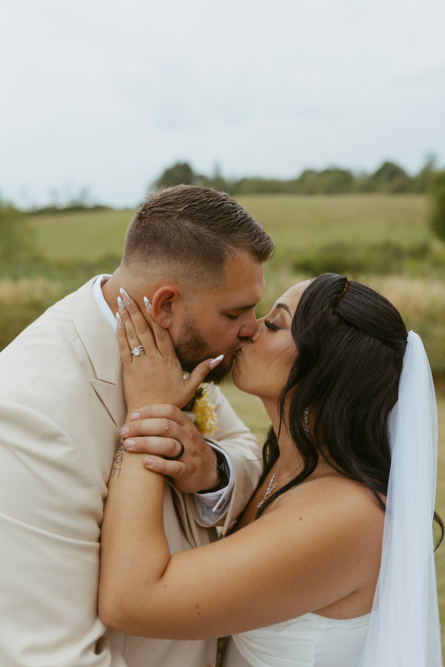 A bride and groom share a romantic kiss outdoors in a field, with the bride wearing a veil and the groom in a beige suit.