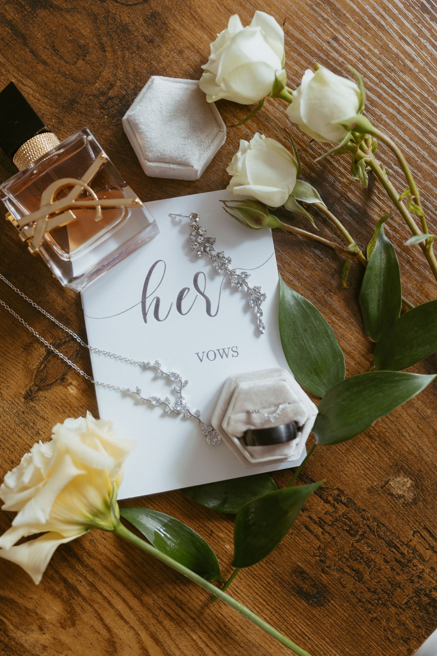 Elegant wedding flat lay with diamond jewelry, vow book, perfume, ring box, and white roses on rustic wood.