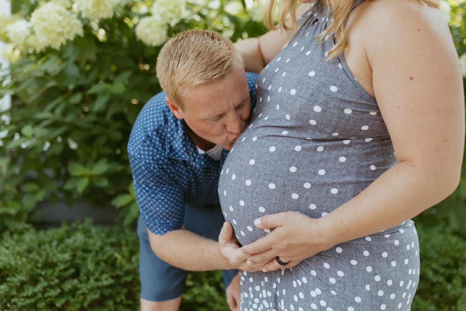 A man kisses a pregnant woman's belly outdoors during a maternity photo session in a lush green garden.