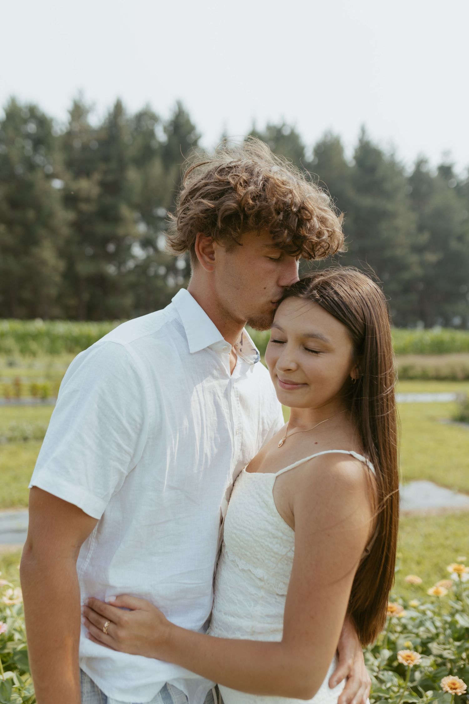 A young couple embraces outdoors, man kissing woman's forehead, both in white attire with green trees behind.