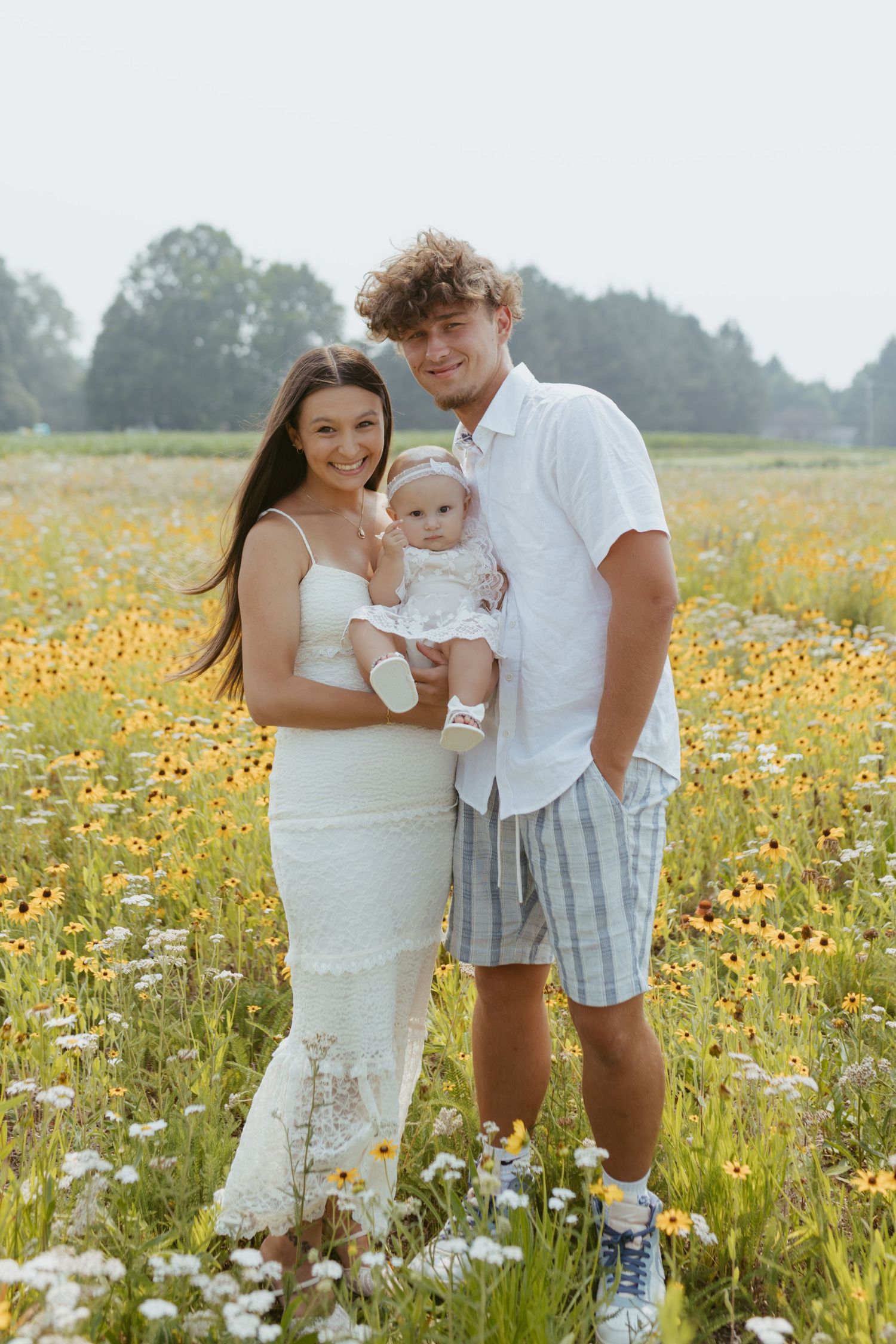 Young family with baby posing in a vibrant wildflower field with yellow and white blooms on a sunny day.