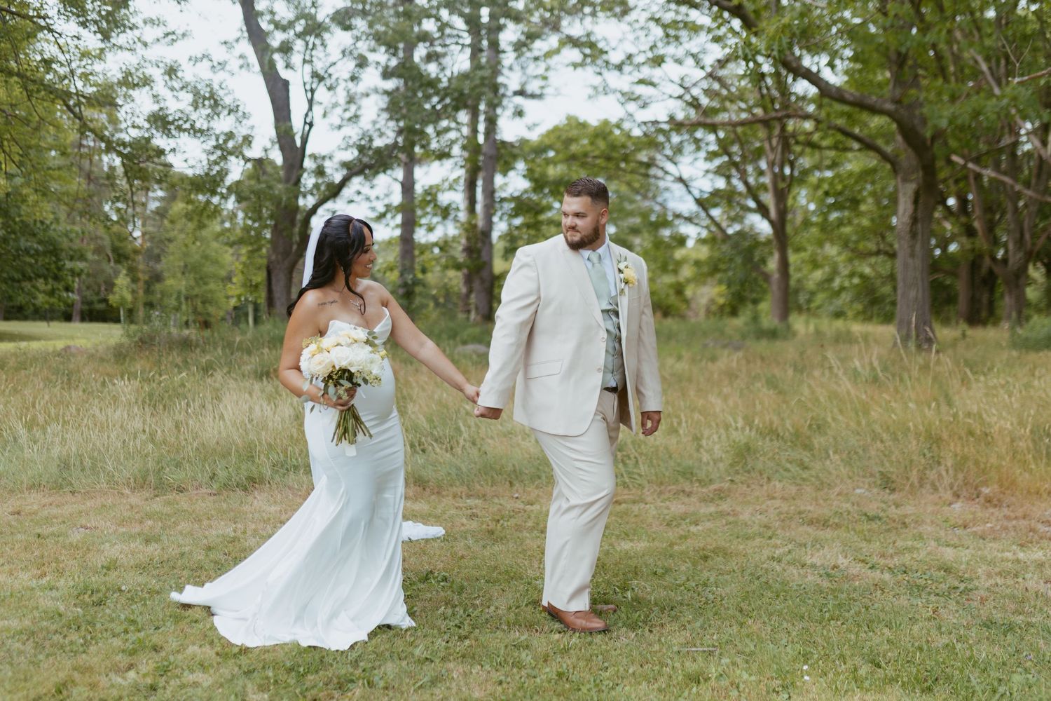 Bride in white gown and groom in tan suit hold hands walking through a lush green field on their wedding day.