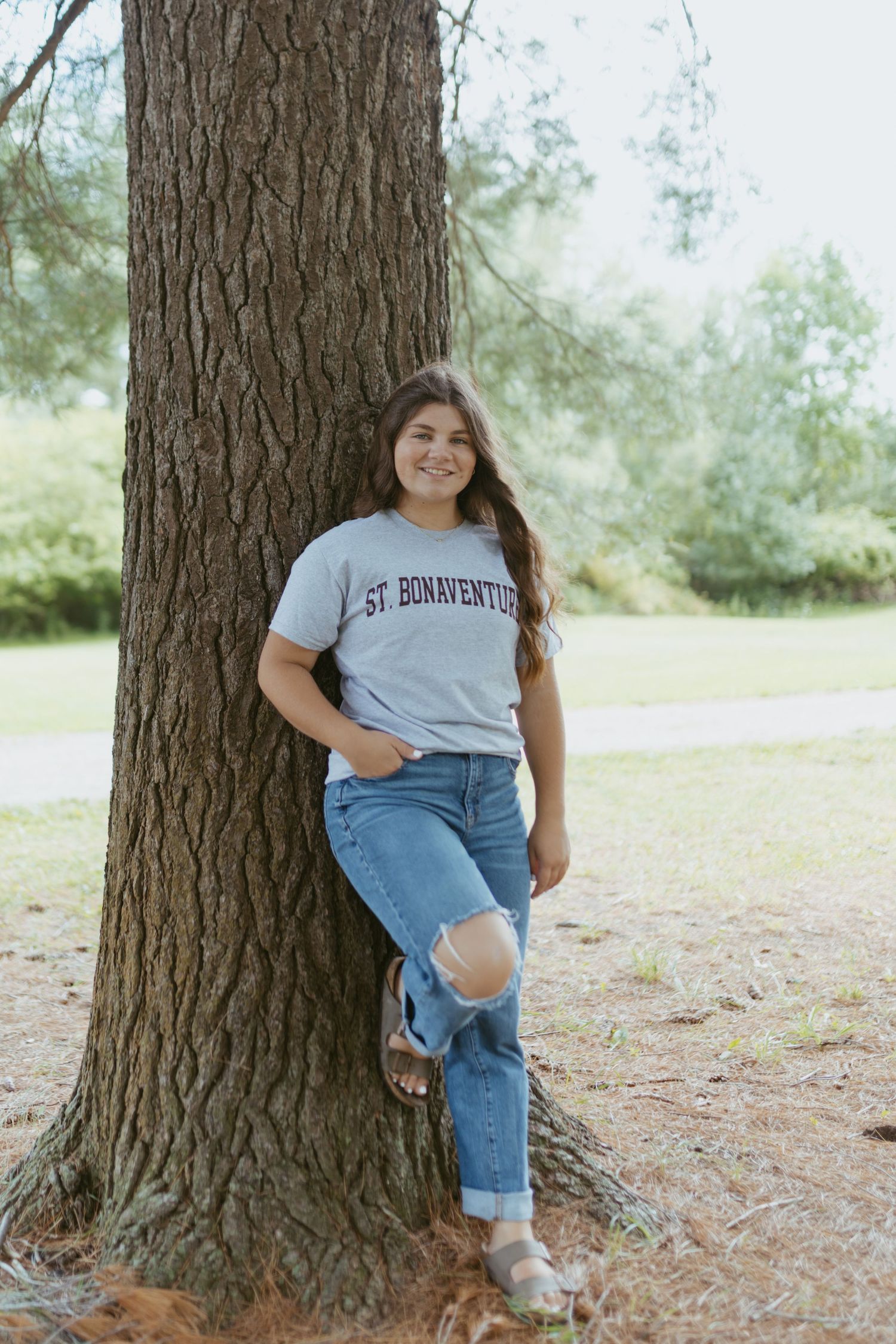 Young woman in St. Bonaventure shirt and ripped jeans leaning against a large tree in a park setting.