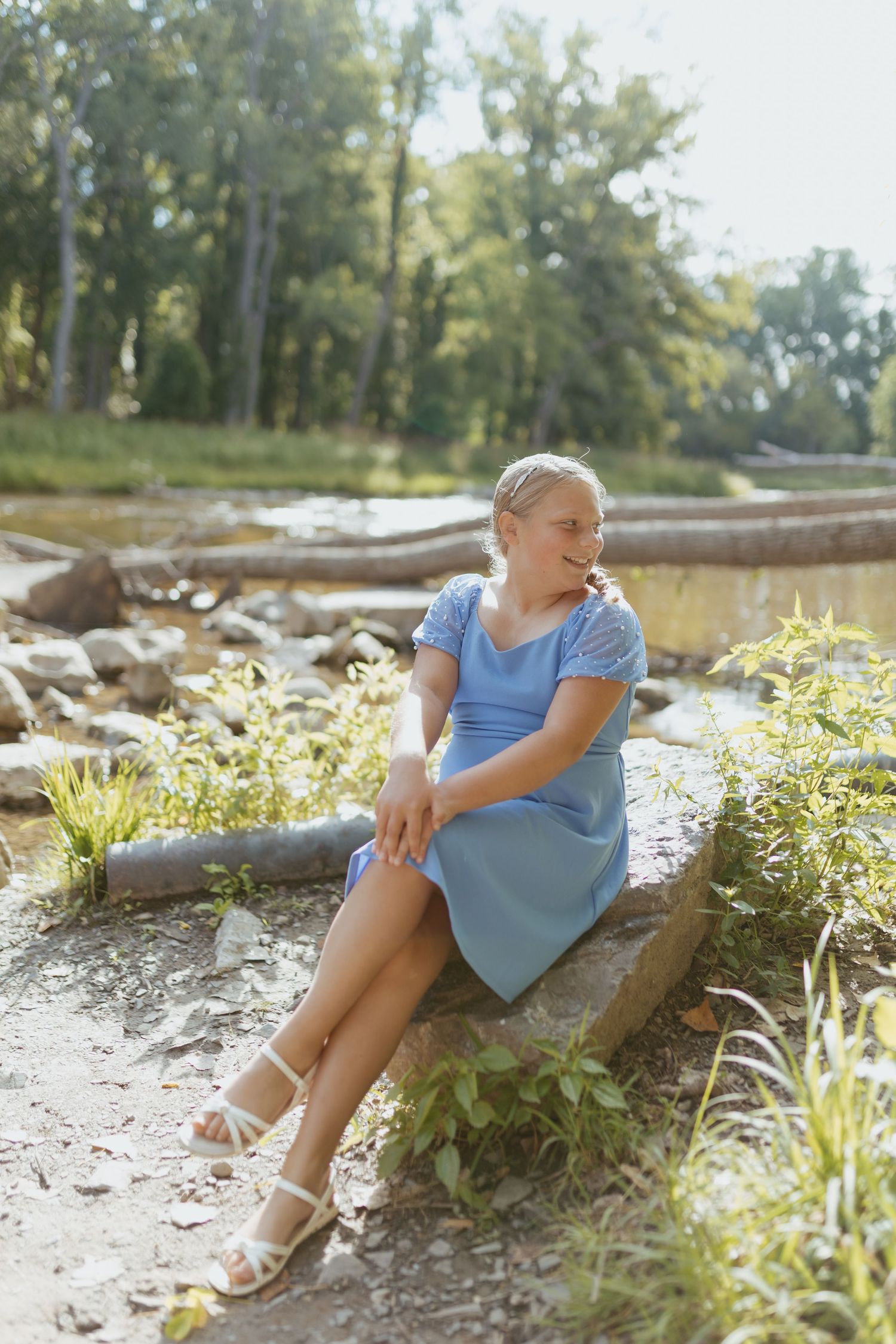 Young woman in blue dress sitting on a rock in a sunny park setting with trees and natural scenery.